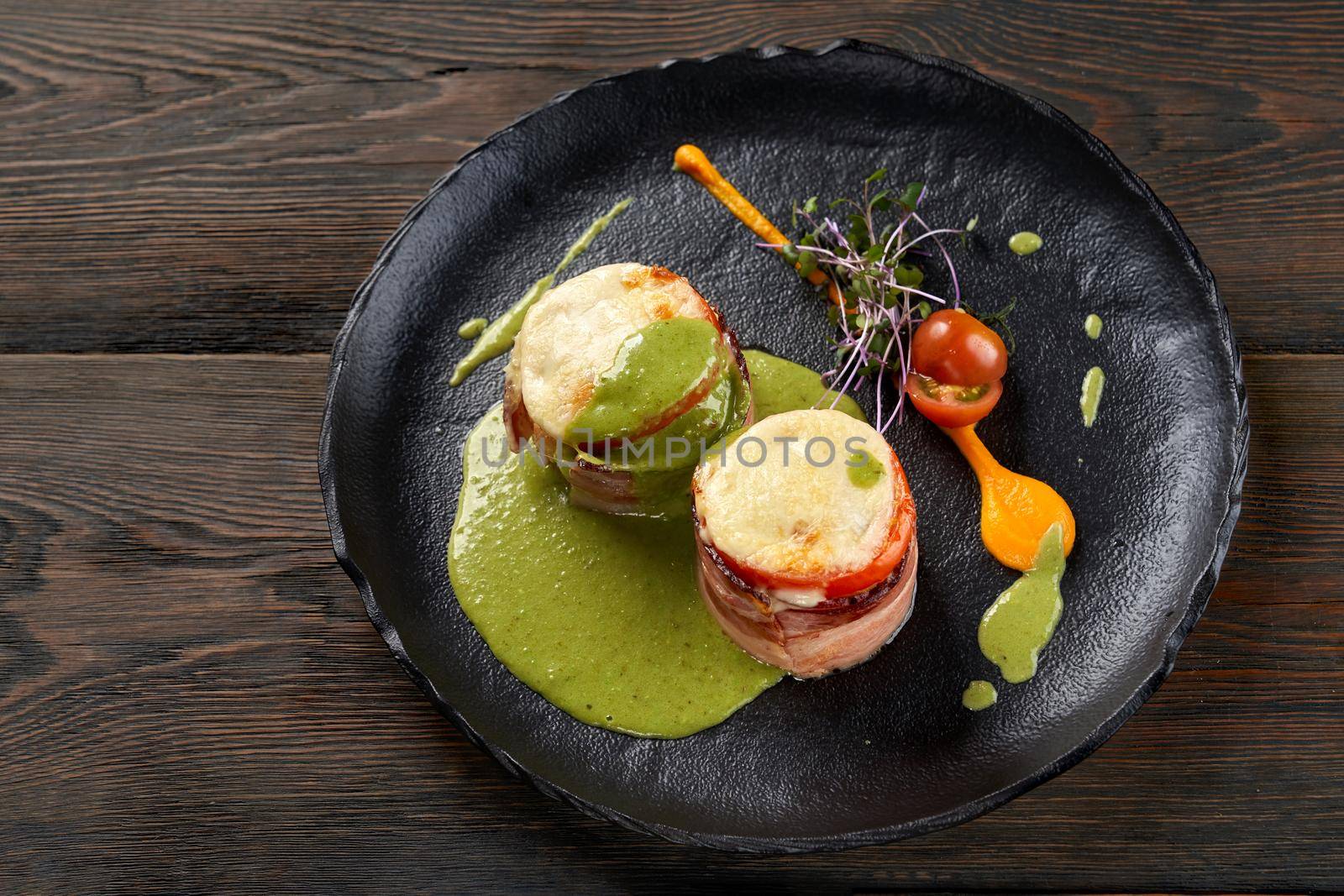 From above view of healthy baked vegetables and ham sandwiches with cheese on top served with pesto sauce, egg yolk, fresh sprouts and cherry tomatoes. Black plate on wooden table.