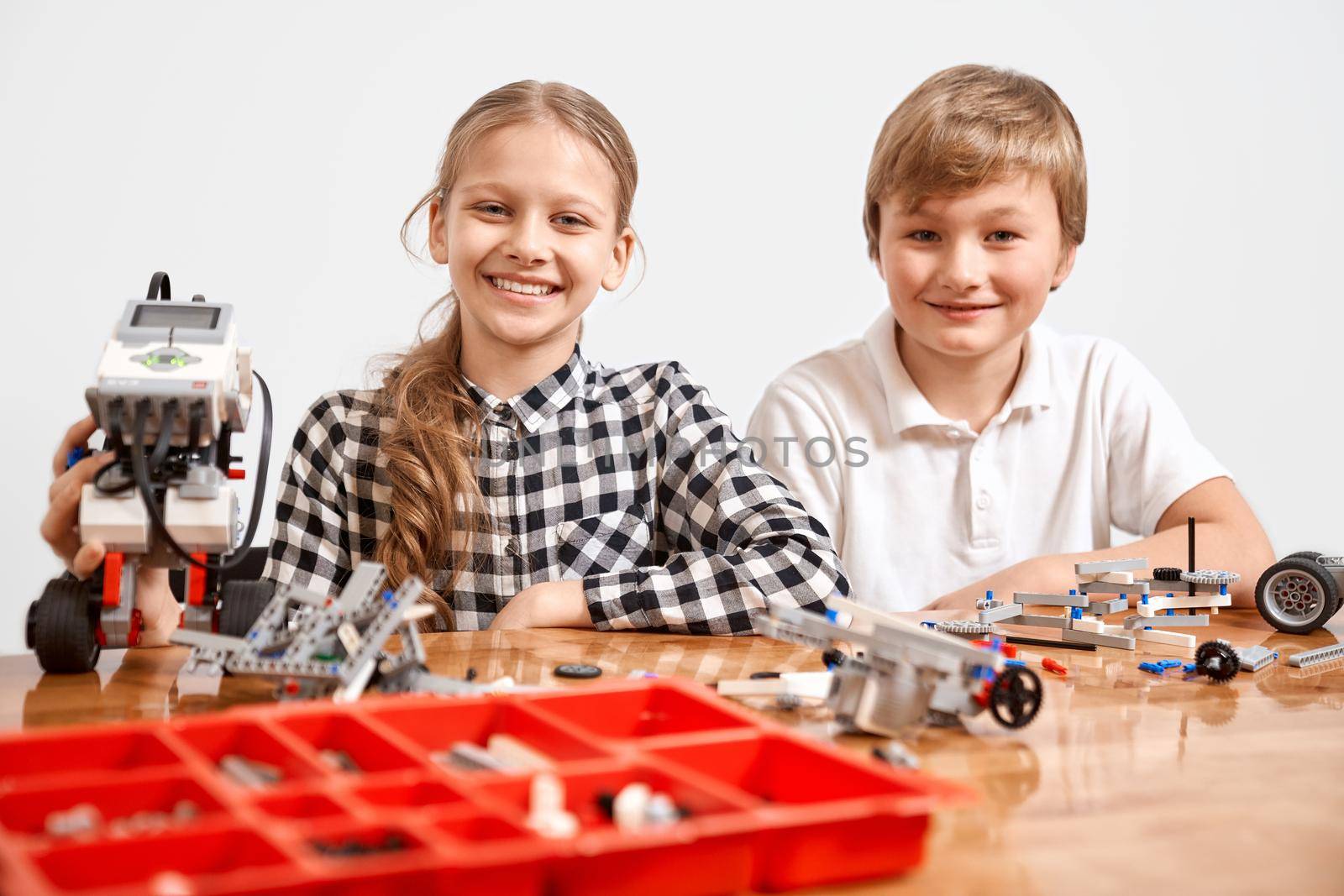 Front view of boy and girl creating robot, red box with building kit on table. Nice interested friends smiling, lookig at camera and working on project together. Concept of science engineering.