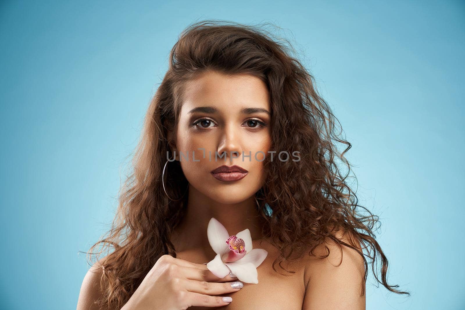 Headshot of young beautiful brunette female model holding orchid on naked chest and looking at camera. Pretty woman with curly hair posing holding pink flower in hand. Beauty, modeling concept.