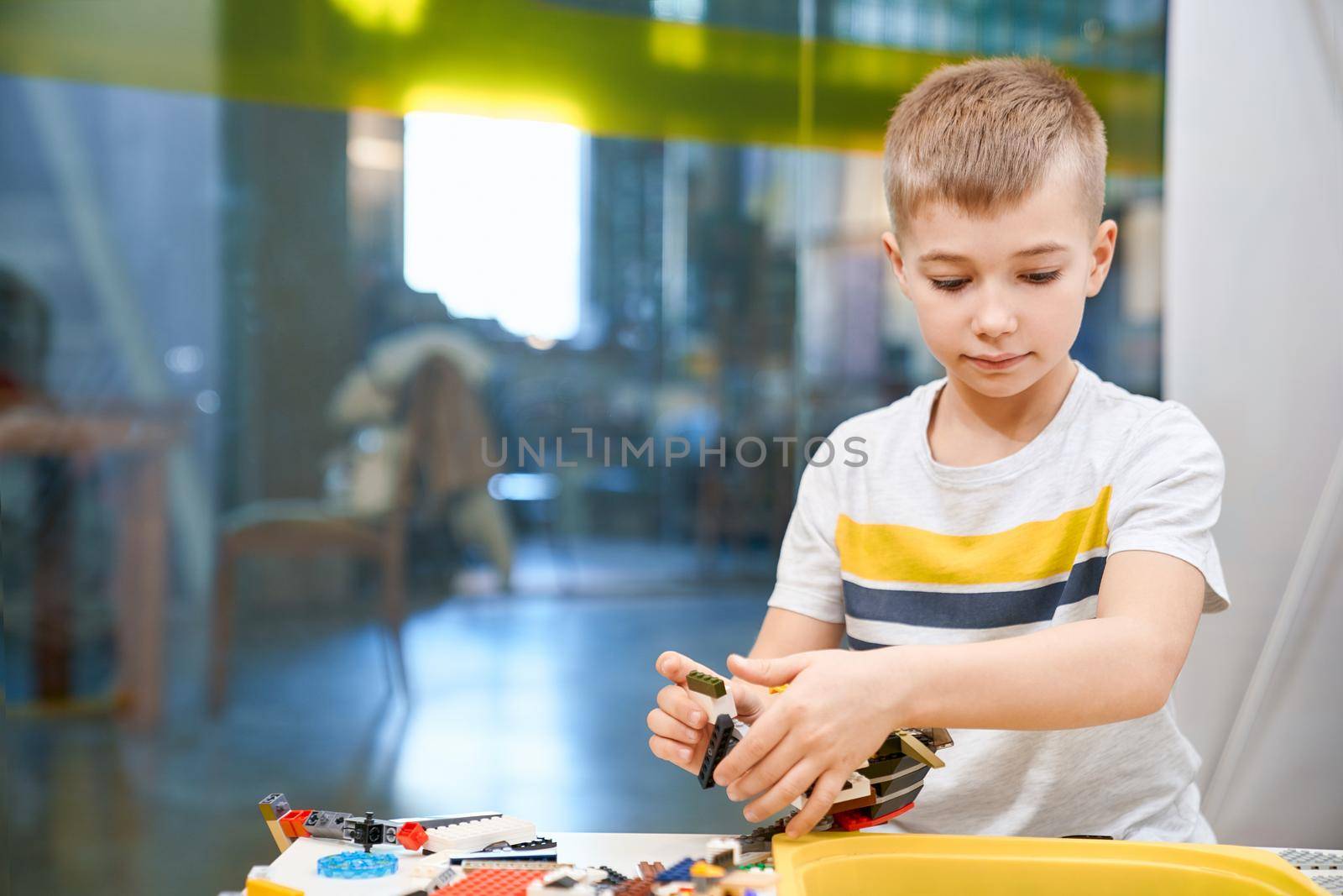 Selective focus of lovely caucasian boy working on project, taking colorful parts to create robot, having positive emotions and joy. Front view of building kit for kids. Science engineering.