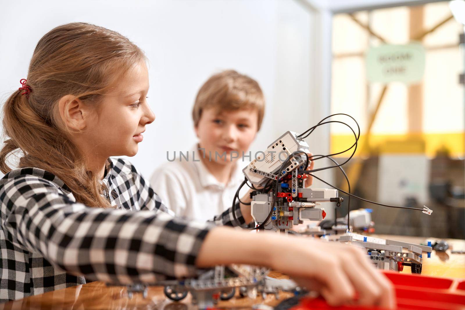 Side view of boy helping girl in creating robot using building kit for kids on table. Science engineering. Nice interested friends smiling, chatting and working on project together.