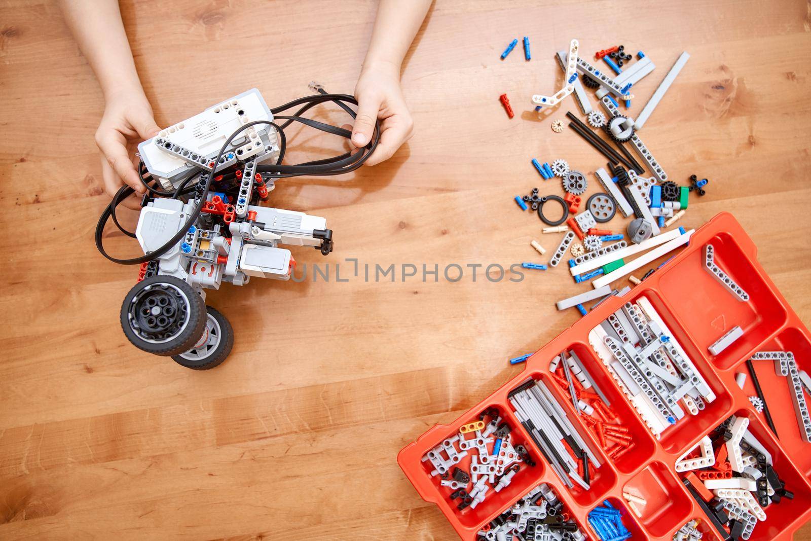 From above view of hands of incognito kid holding robot and working on project, details in box. Colorful pieces from building kit for children on table. Concept of science engineering.