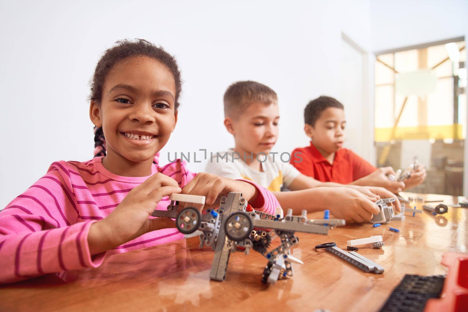 Side view of building kit for group of three multiracial kids creating toys, having positive emotions and joy, african girl smiling and looking at camera. Close up of friends working on project.