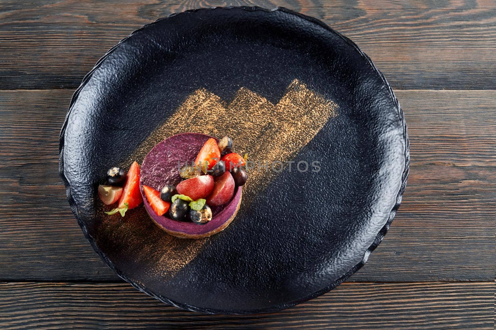 From above view of round piece of traditional blueberry cheesecake served with blueberries and strawberries on black plate on wooden table in restaurant, golden brushstroke. Concept of food serving.
