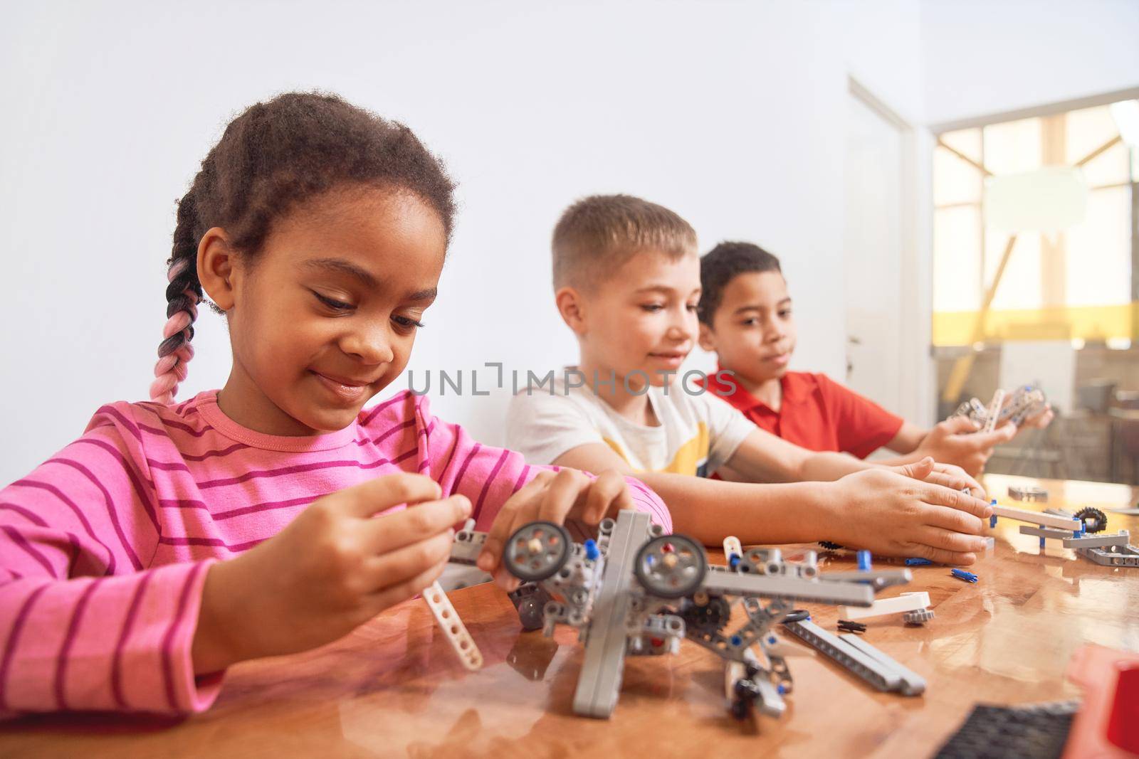 Building kit with colorful pieces for group of three multiracial kids creating toys, having positive emotions. Close up of smiling african girl working on project. Concept of science engineering.