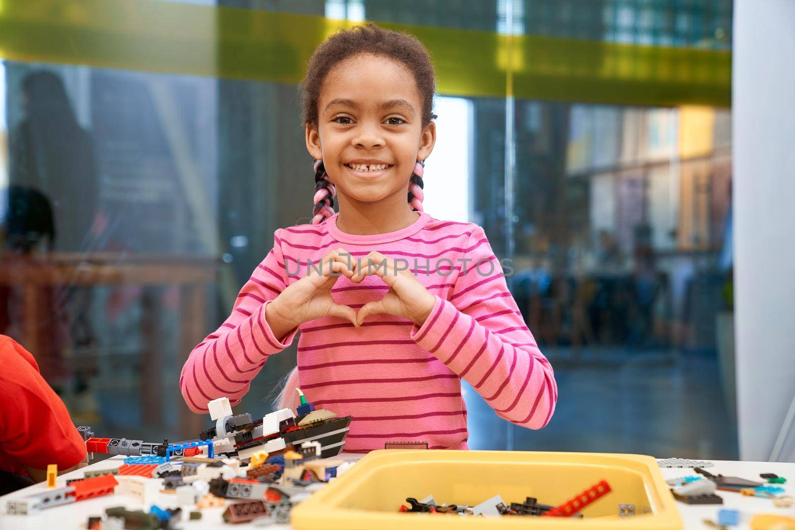 Front view of lovely smiling african girl standing, looking at camera and making heart with hands. Building kit for kids on table, children creating toys, having positive emotions and joy.