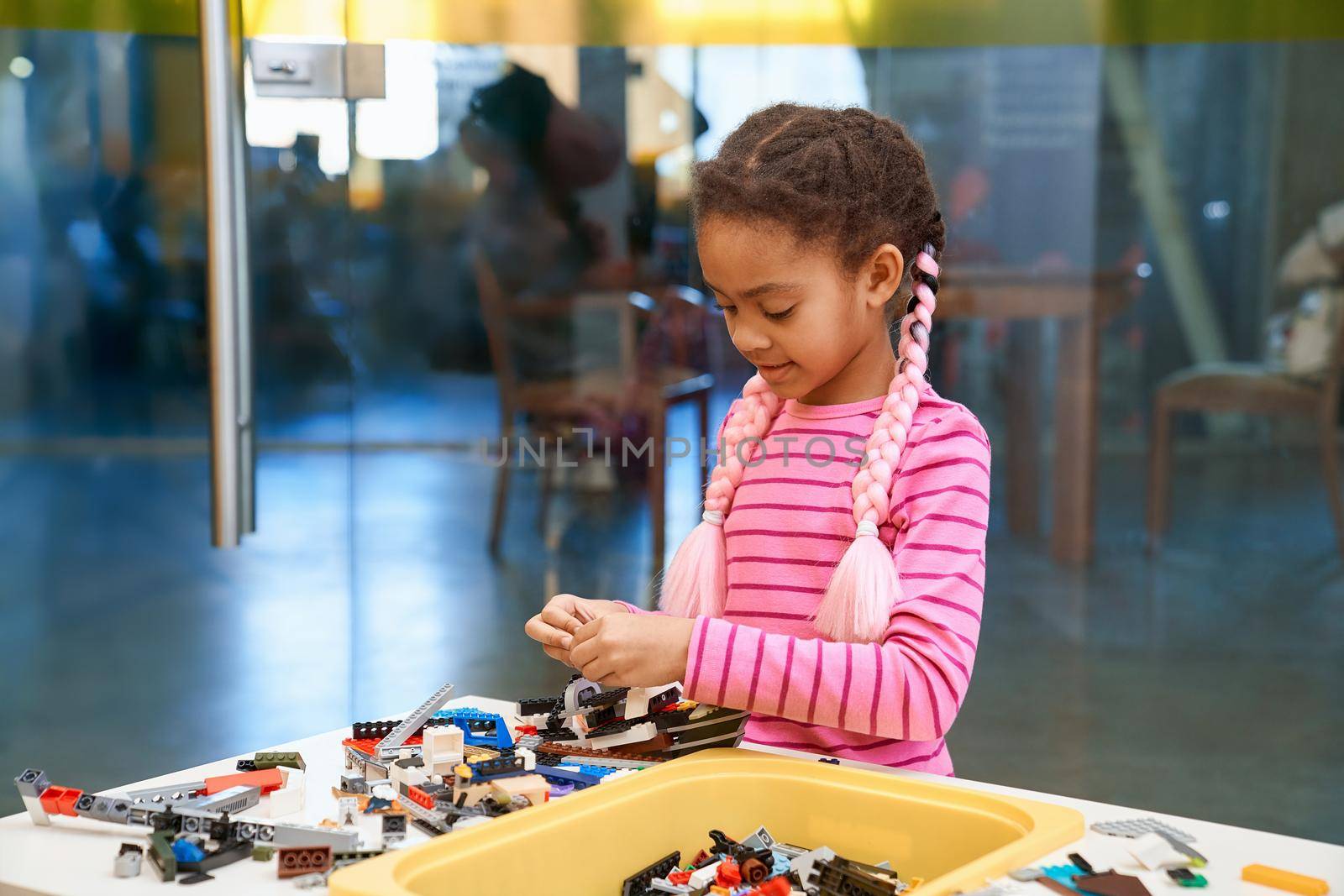 Front view of lovely conentrated african girl standing. Building kit for kids on table, children creating toys, having positive emotions and joy. Close up of kid working on project.