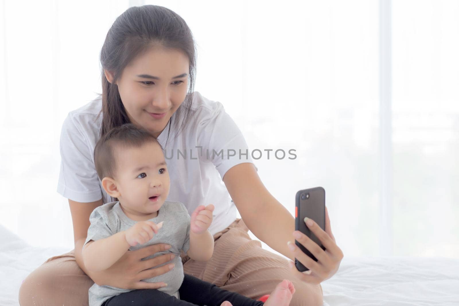 Young asian mother and little baby girl or newborn selfie with smart phone on bed in bedroom, happiness mom and daughter using phone video call at home, two people, family and communication concept.