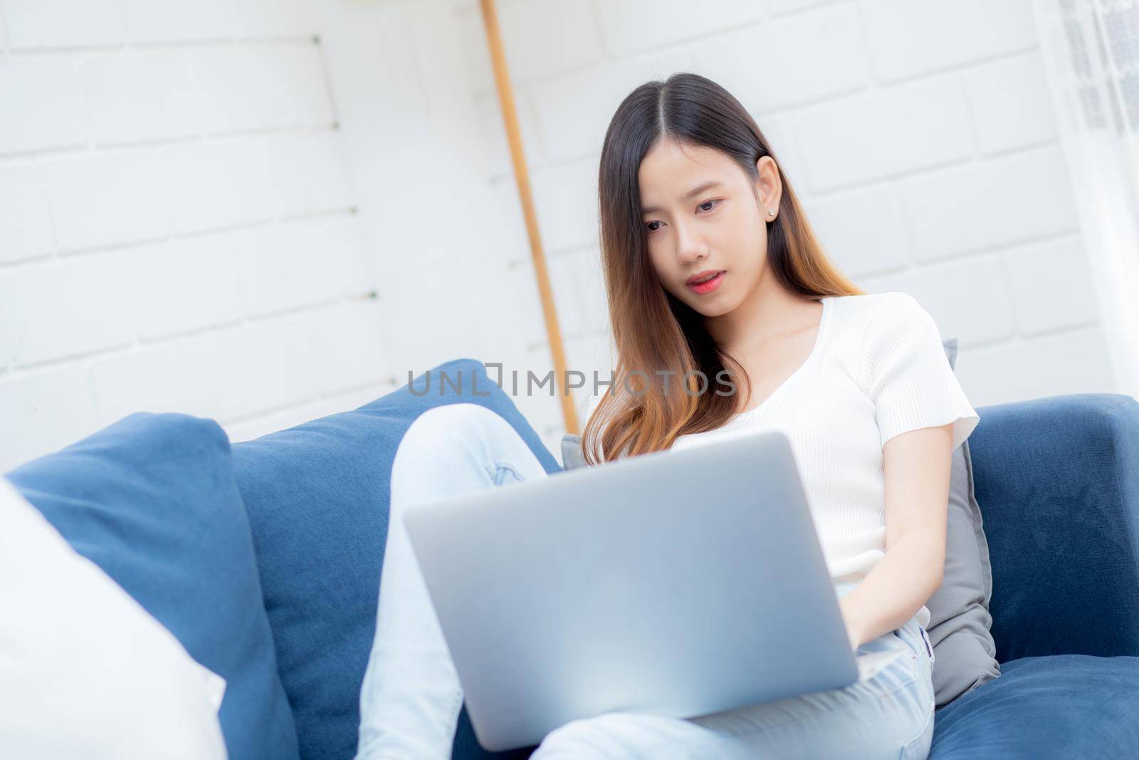 Young asian business woman smile and work from home with laptop computer online to internet on sofa in living room, freelance girl using notebook on couch with comfort, new normal, lifestyle concept.