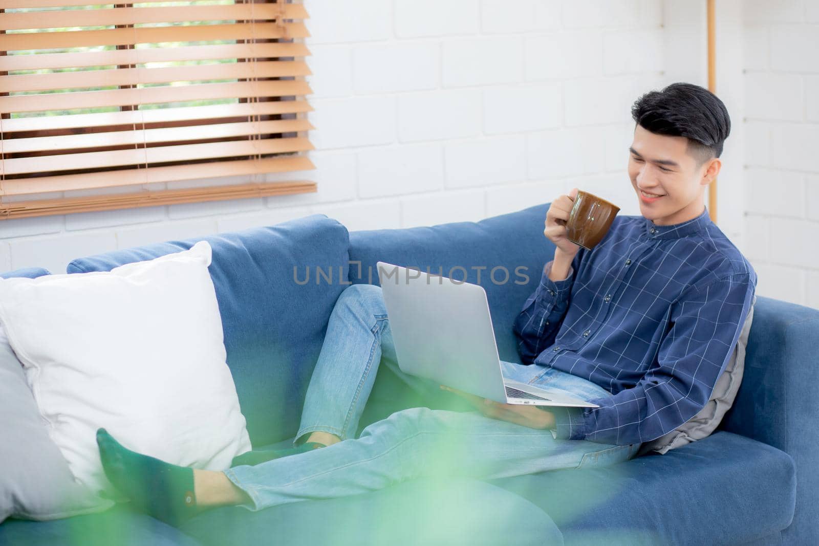 Young asian businessman smile and work from home with laptop computer online on sofa in living room, freelance business man using notebook and drink coffee on couch, new normal, lifestyle concept.