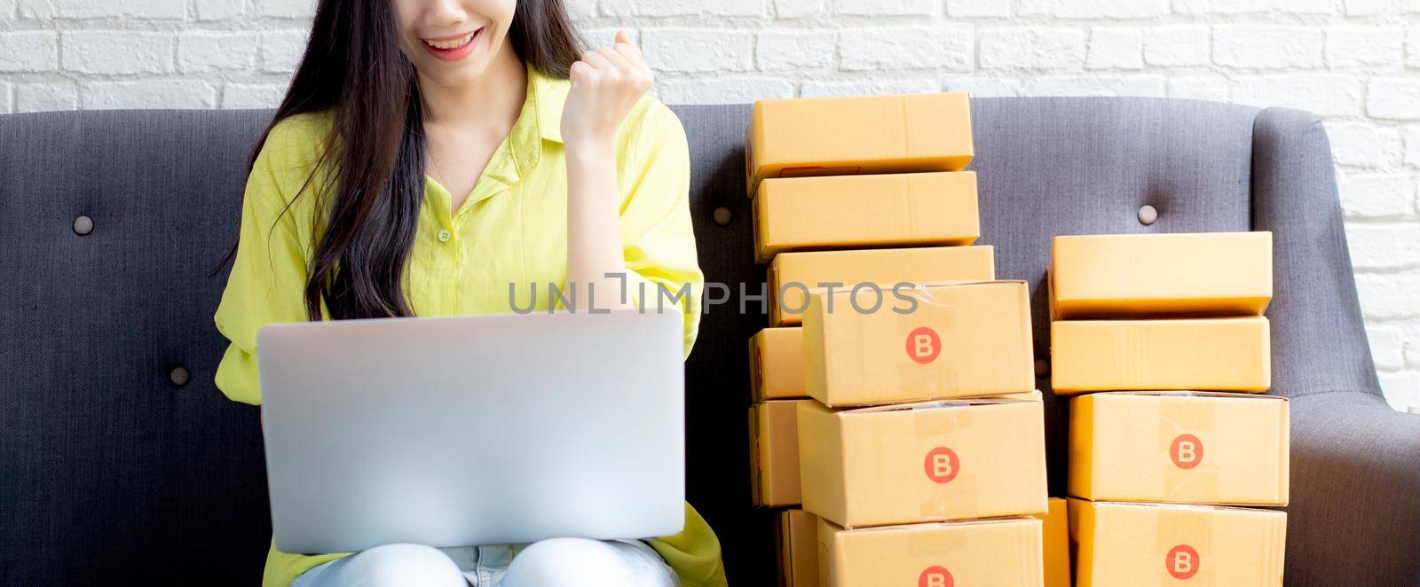 Young asian woman is merchant using laptop computer on sofa checking order of customer with purchase online shopping, business SME or startup, entrepreneur or seller and service delivery product.