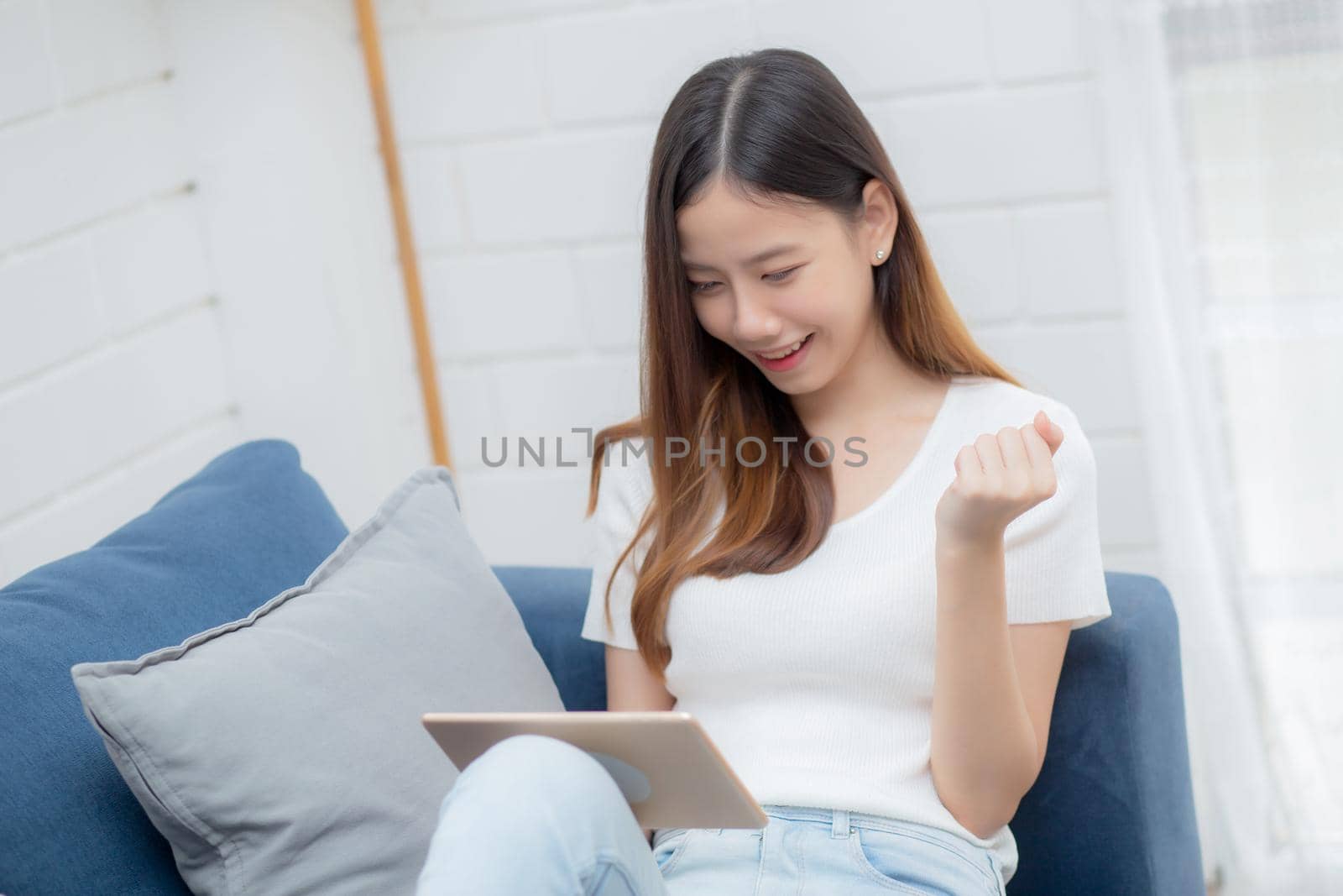 Young attractive asian woman resting using browsing tablet computer on sofa at home, happy girl sitting on couch relax reading digital gadget with excited at house, communication and lifestyle concept.