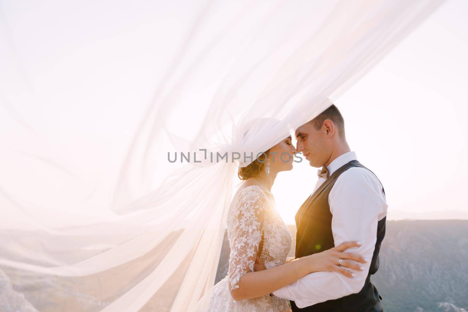 The wedding couple cuddles on top of the mountain at sunset, the groom hugs the bride by the waist, the veil is waved into the air.