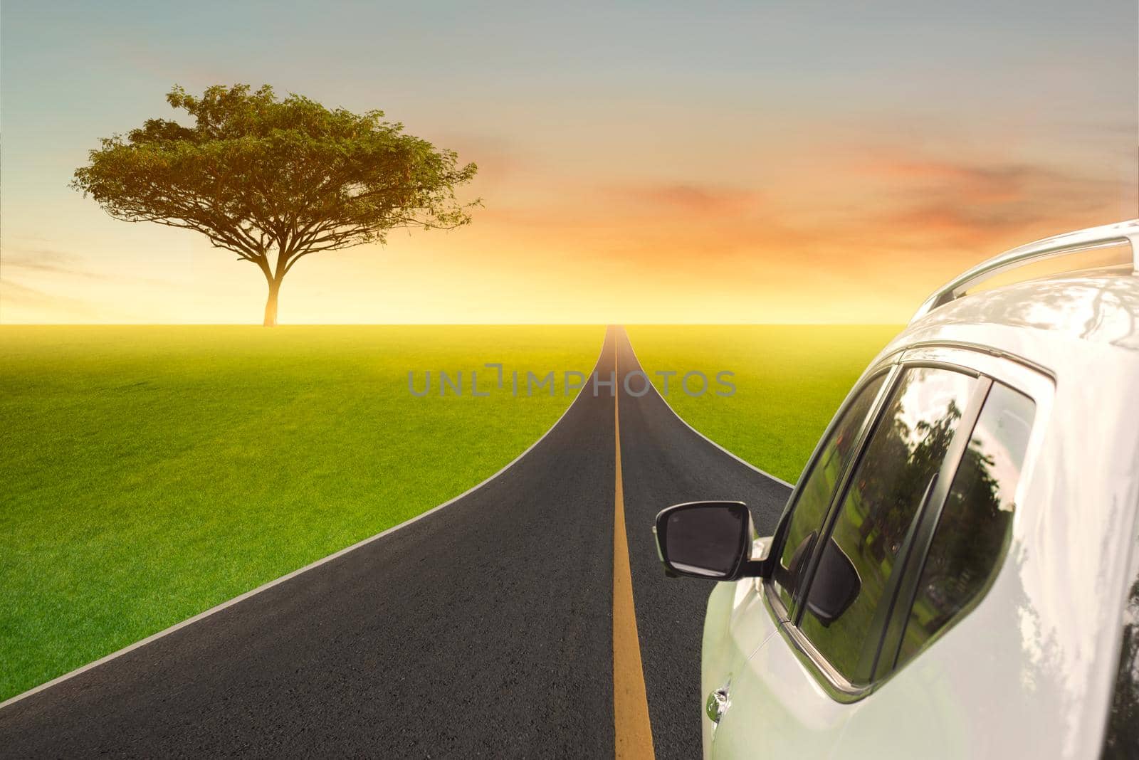 A white car driving on the countryside asphalt road against green field with clouds and a beautiful sunset.