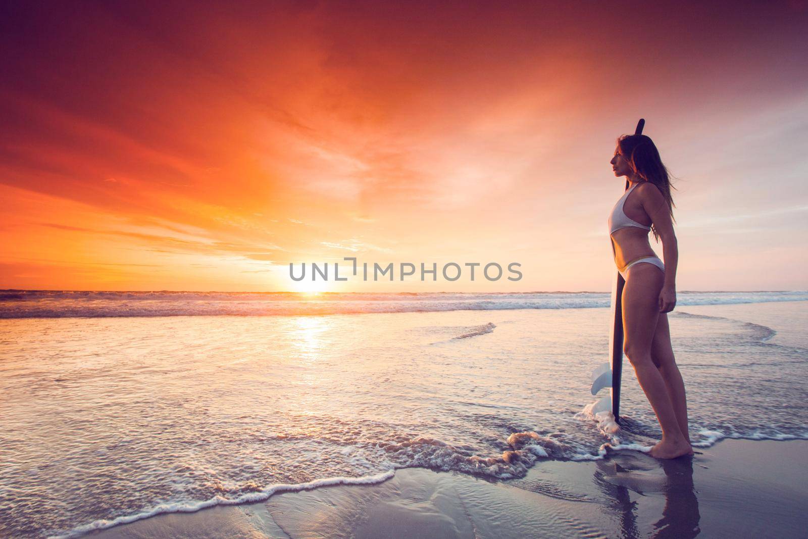 Woman holding surfboard standing disappoint in the edge of sea wave, looking forward to the sea for sufficeint wave to surf at sunset on background