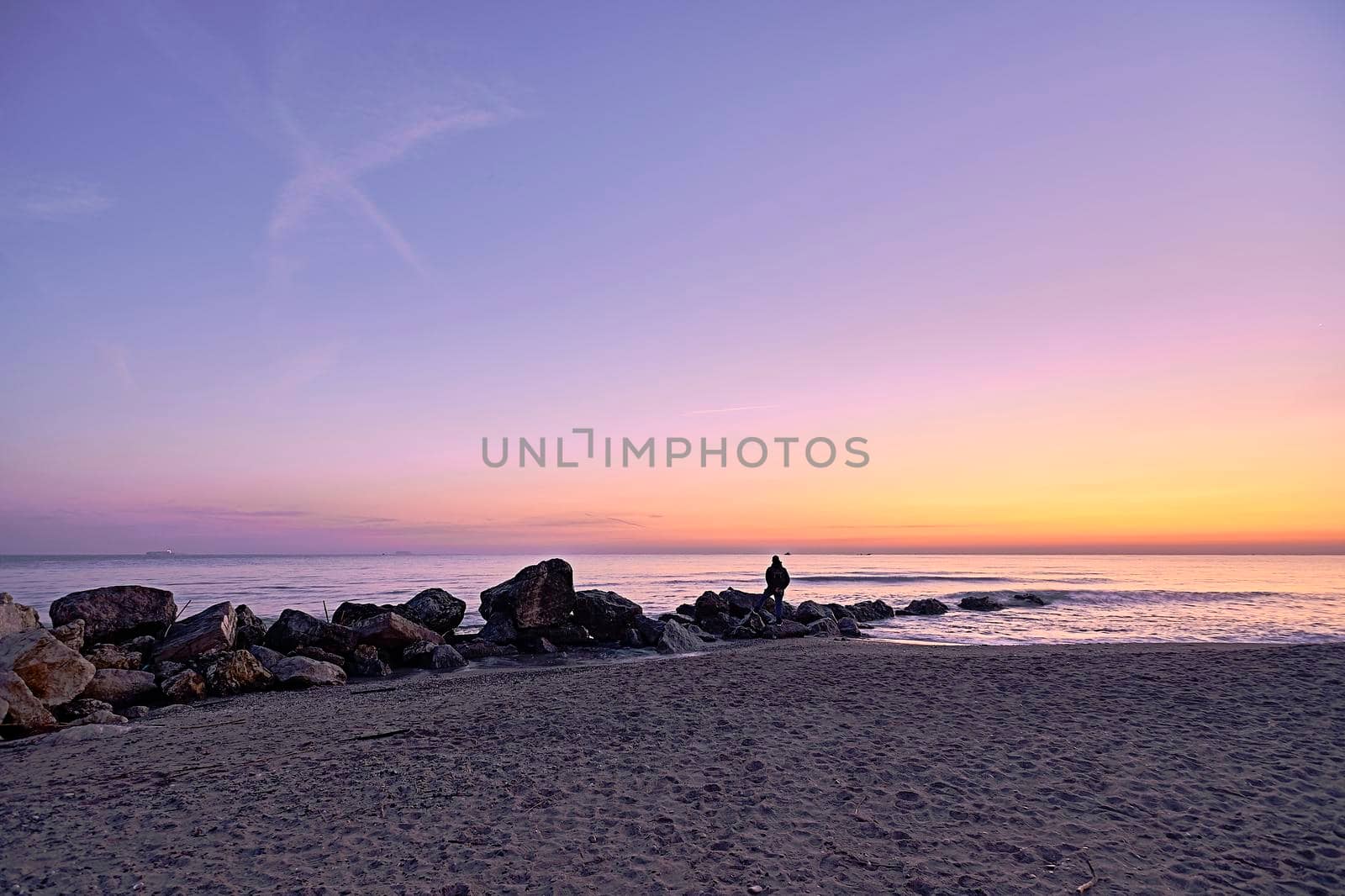 Man watching the sunrise from the rocks on the beach, lonely, orange colors, sand, calm sea