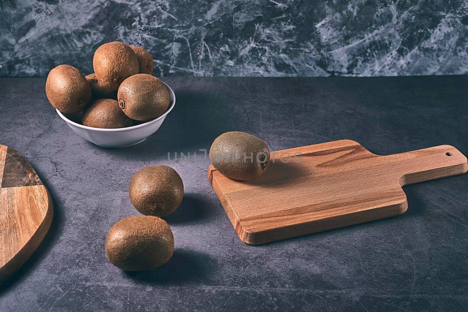 Three kiwis on a wooden board and one in a bowl. Dark stone floor and marble wall, white bowl.