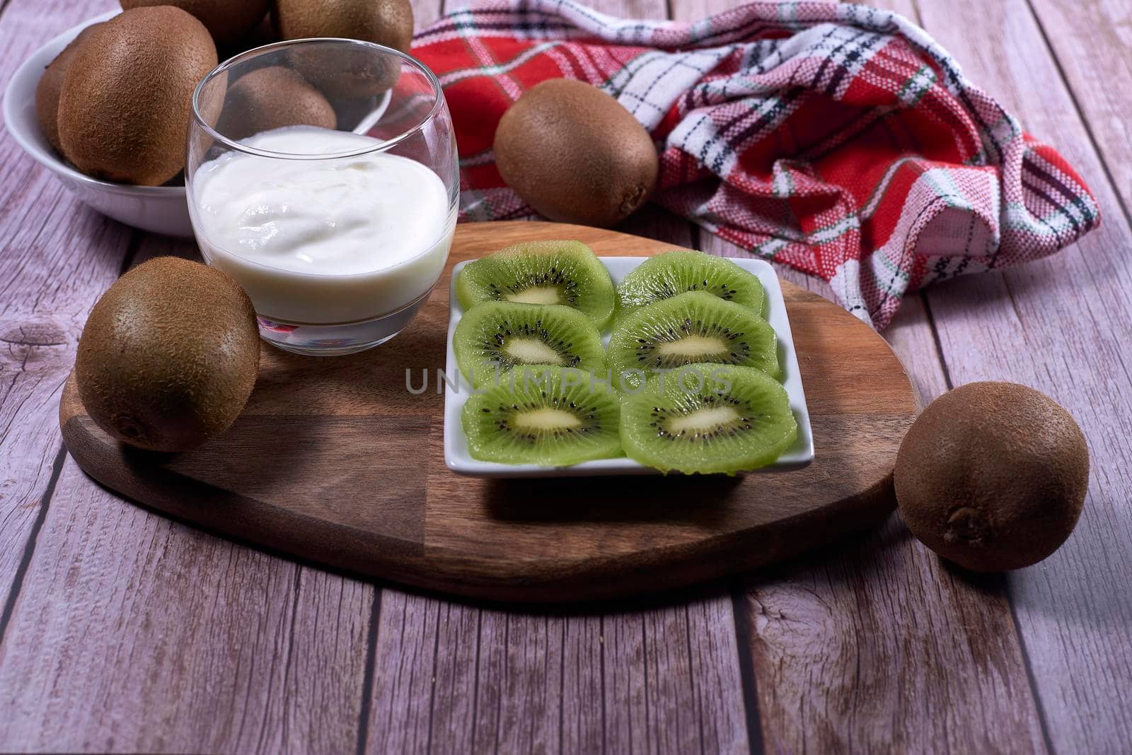 Yoghurt glass and plate of cut kiwis and several whole ones on wood. Wooden board, red and white kitchen towel, squares, white bowl, front view.