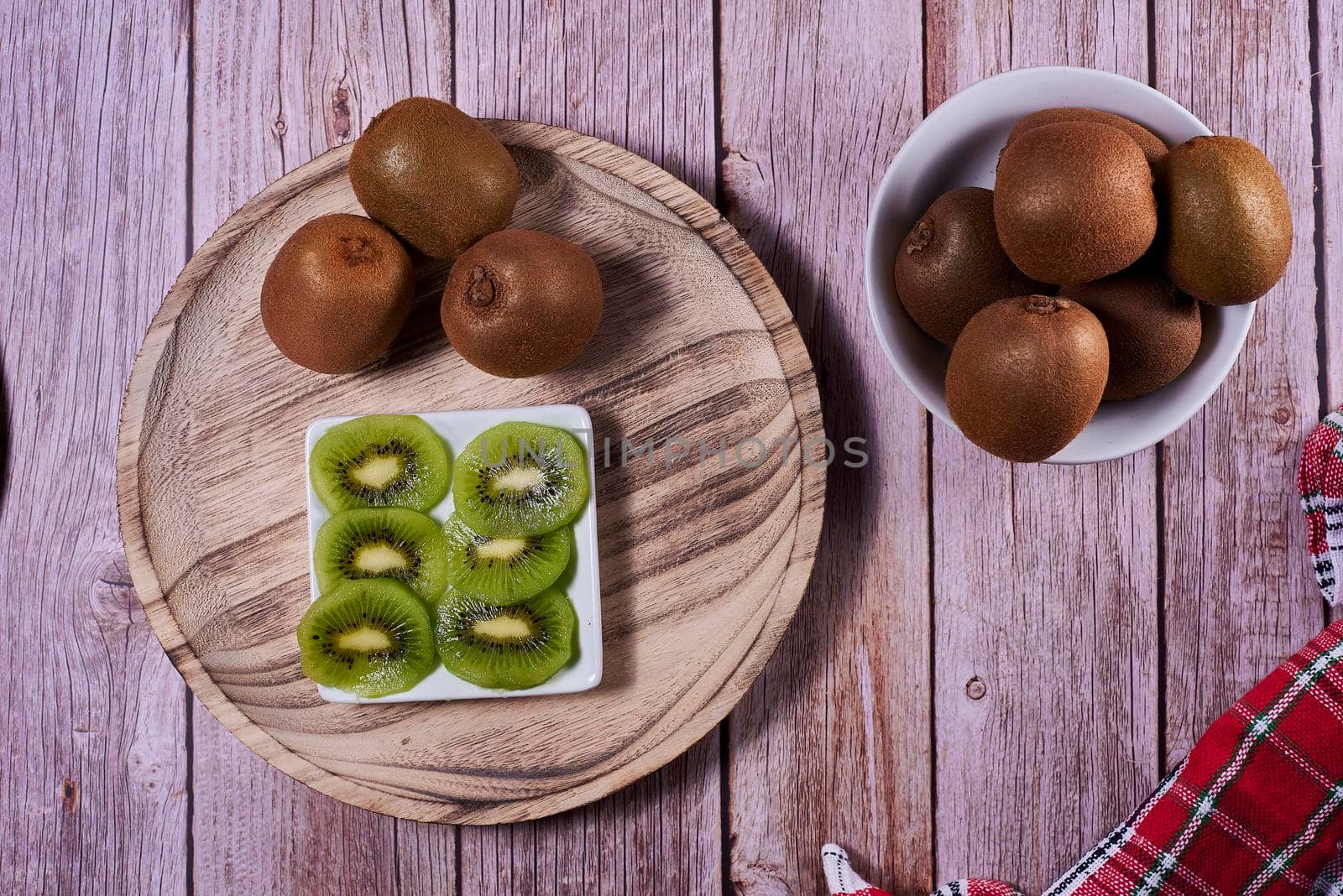 Yoghurt glass with kiwis on wooden plate. Wooden floor, red and white kitchen towel, chequered, zenithal view, kiwi cut on rectangular plate.