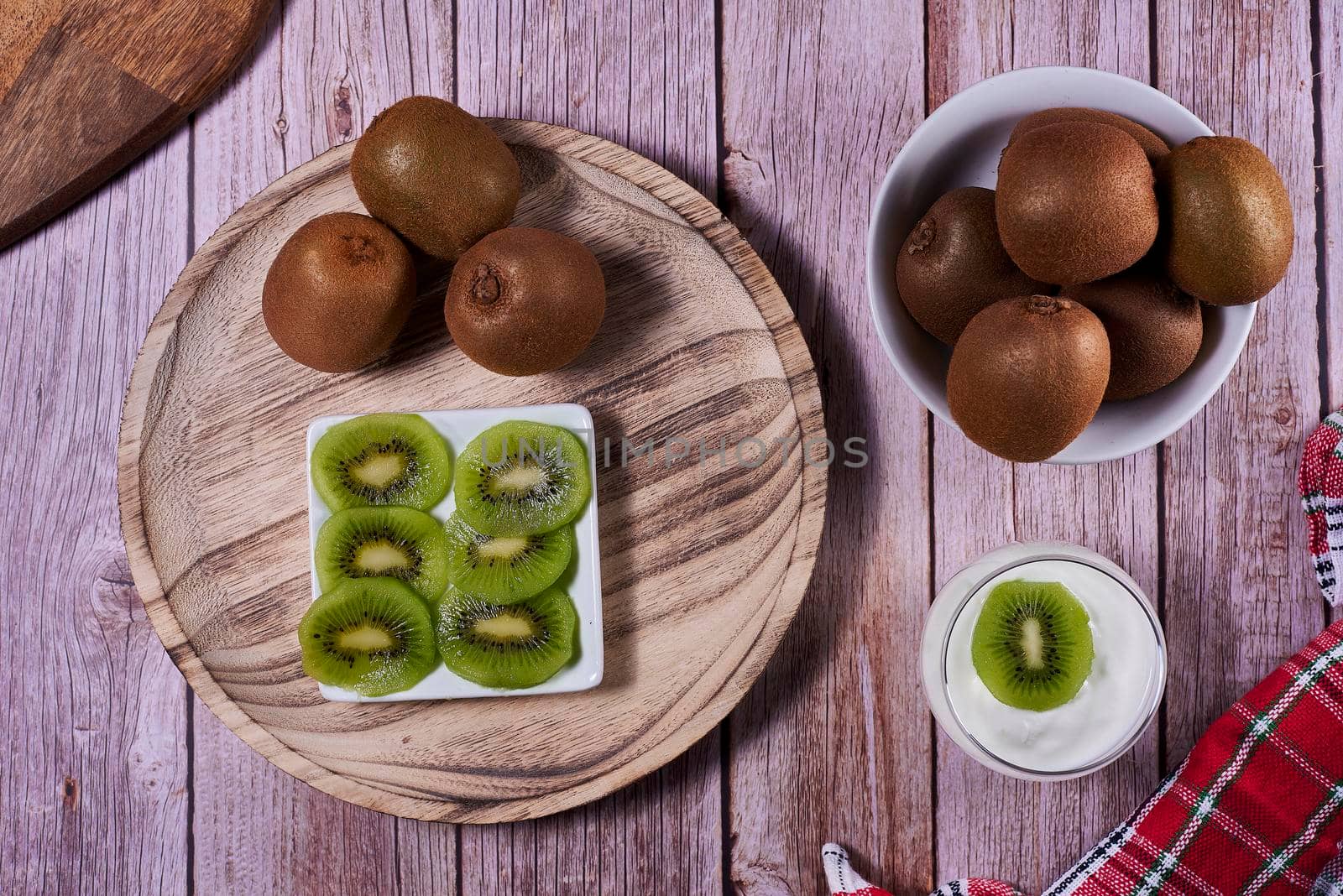 Yoghurt glass with kiwis on wooden plate. Wooden floor, red and white kitchen towel, chequered, zenithal view, kiwi cut on rectangular plate.