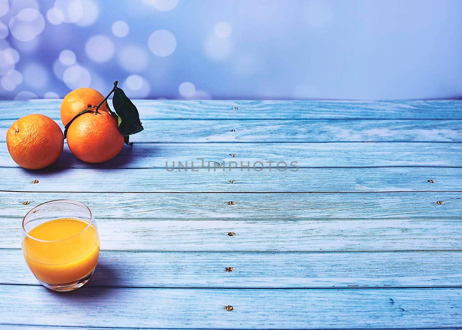 Glass of orange juice on wooden floor with several orange trees and bright background, empty space