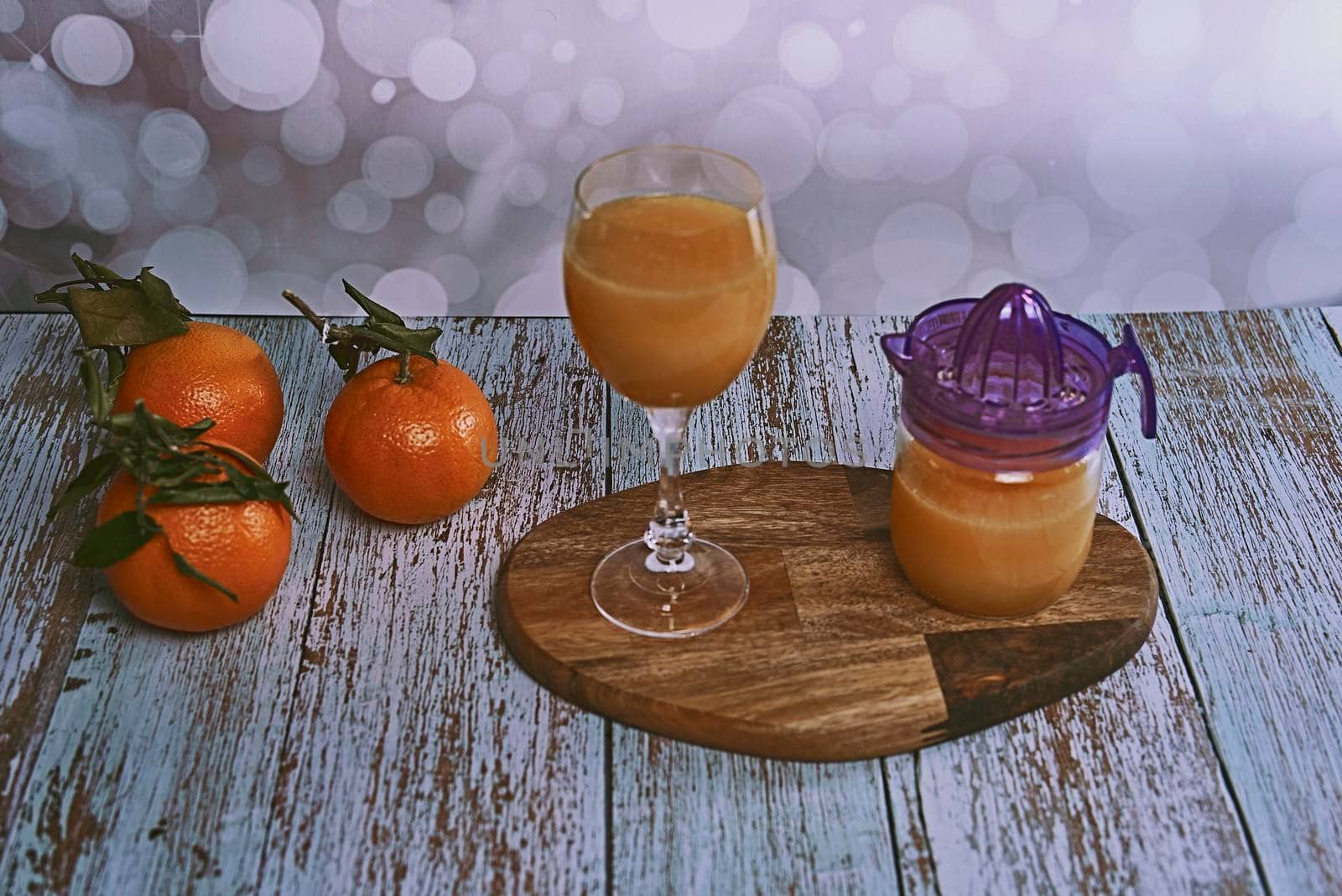 Glass of orange juice with glass juicer on wooden table, various oranges on wooden floor and bright background