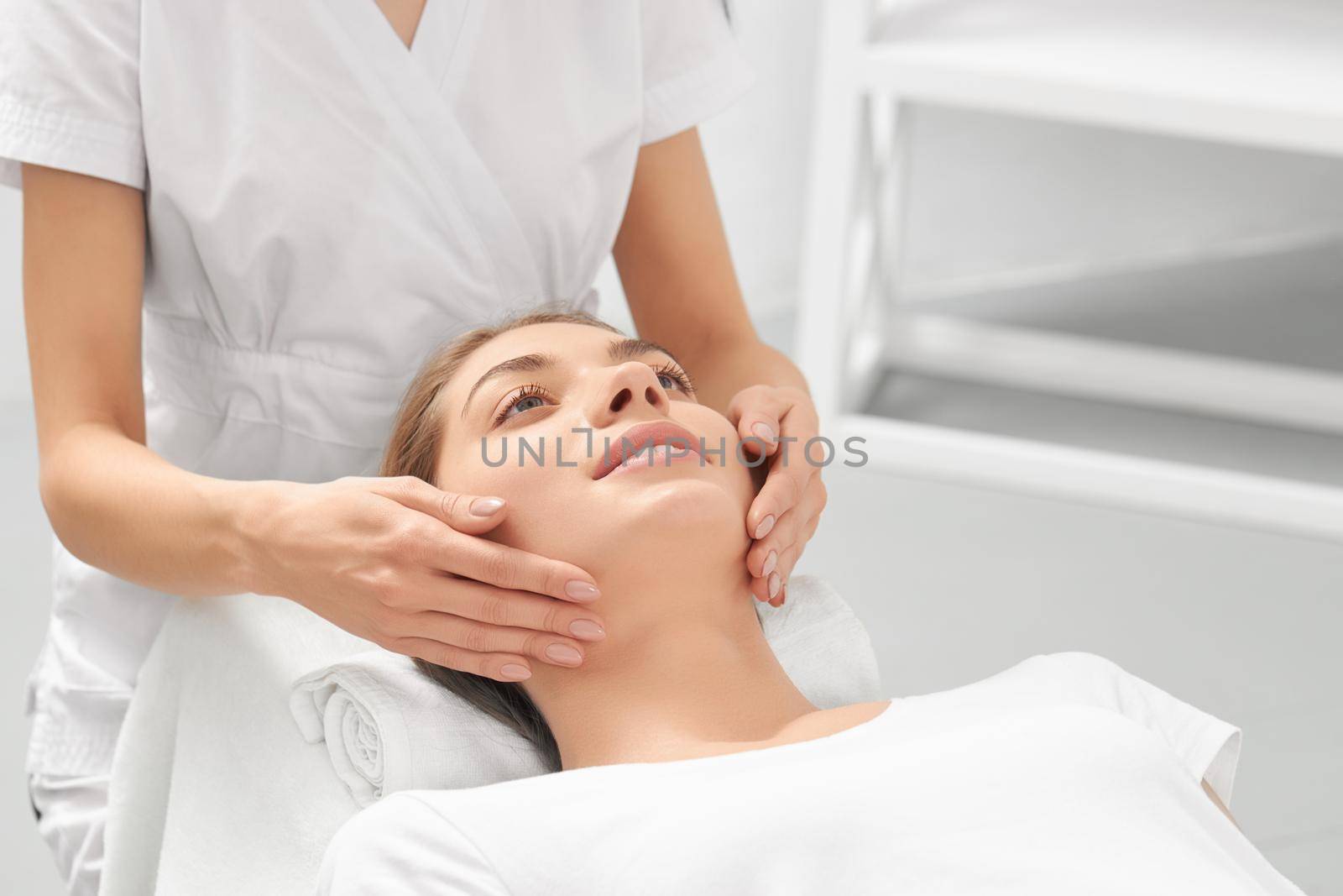 Close up portrait of beautician in white uniform doing massage hand for young smiling woman in professional salon. Concept of process special procedure for improvements skin face and body. 