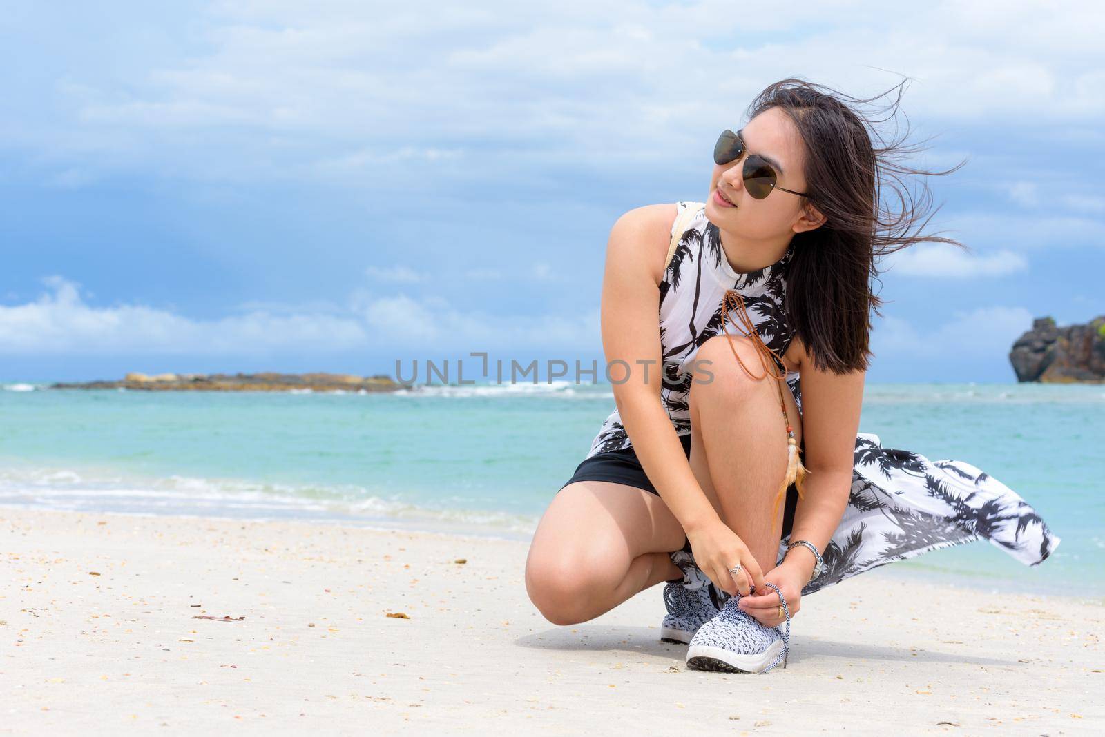 Beautiful woman tourist wearing sunglasse squatting binding rope sneaker on the beach near the sea during the stroll on Tarutao island in the summer vacation, Satun, Thailand