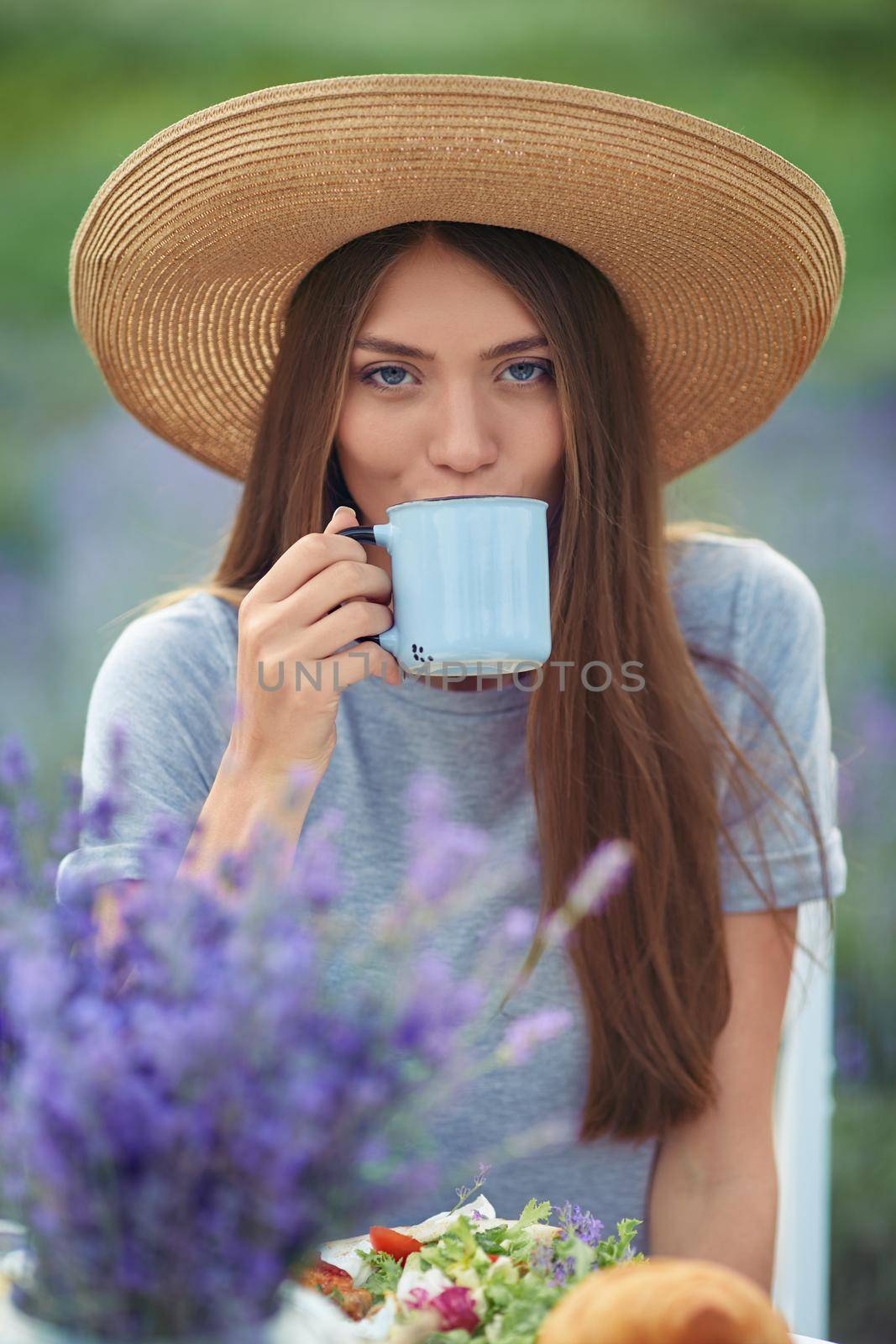 Front view of young woman drinking hot coffee, posing at table served with food and flower bouquet. Portrait of stunning girl wearing straw and dress hat sitting in lavender field, looking at camera.