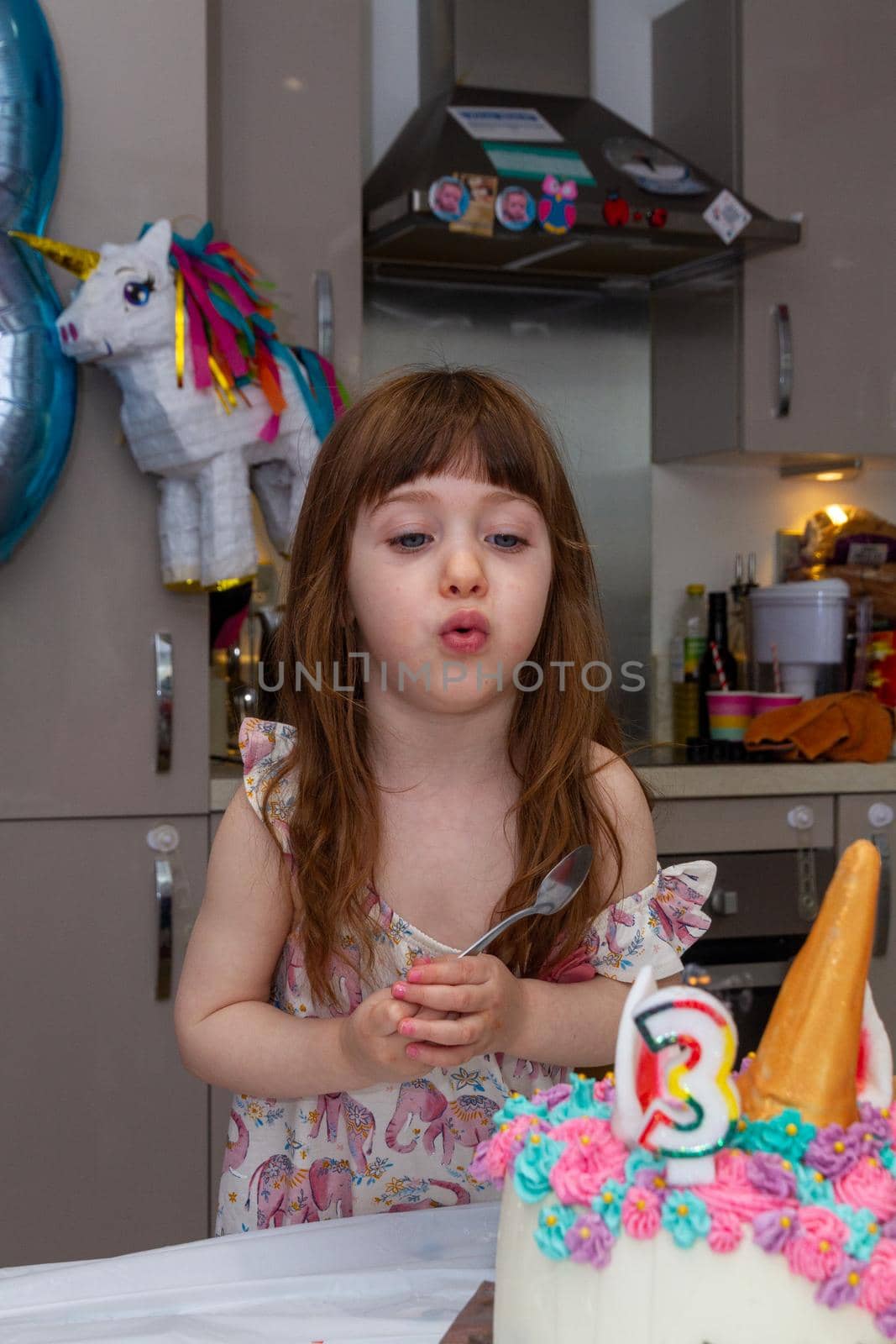 A Cute, brown-haired baby girl in a kitchen blowing candles of a birthday cake decorated as an unicorn with a number three candle with a pinhata in the background