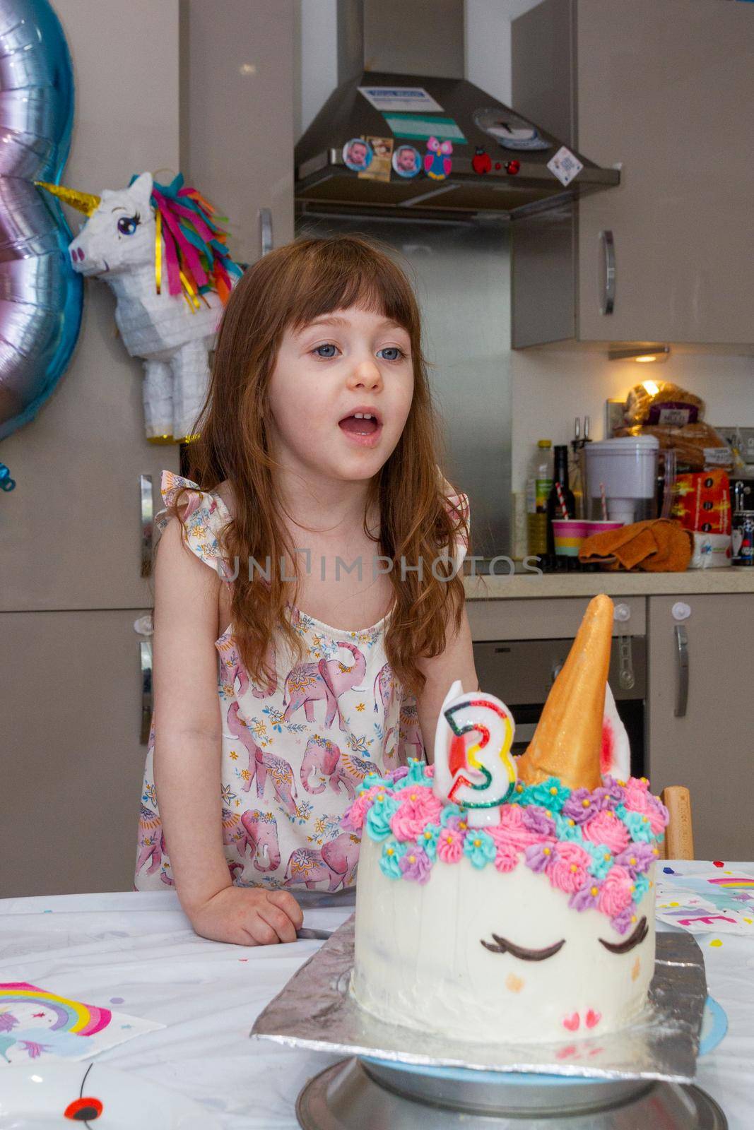 A Cute, brown-haired baby girl in a kitchen blowing candles of a birthday cake decorated as an unicorn with a number three candle with a pinhata in the background