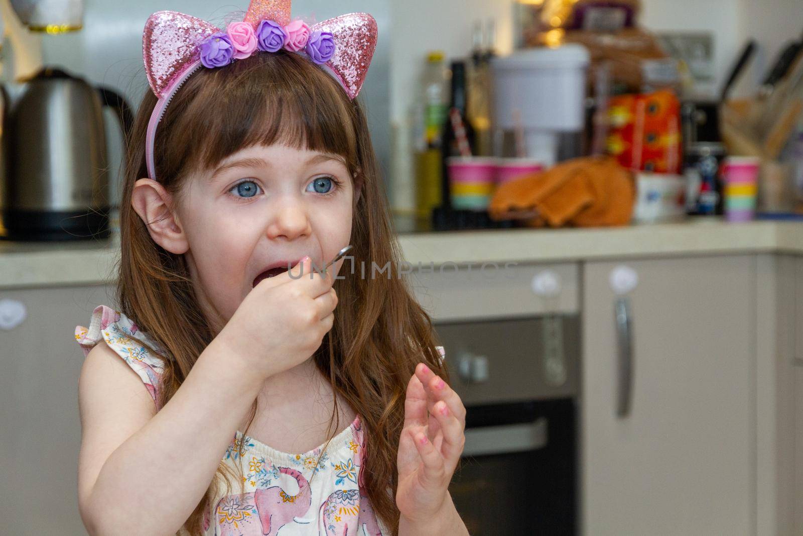 Portrait of a cute, brown-haired, blue-eyed baby girl in a kitchen wearing a headband and eating with a spoon