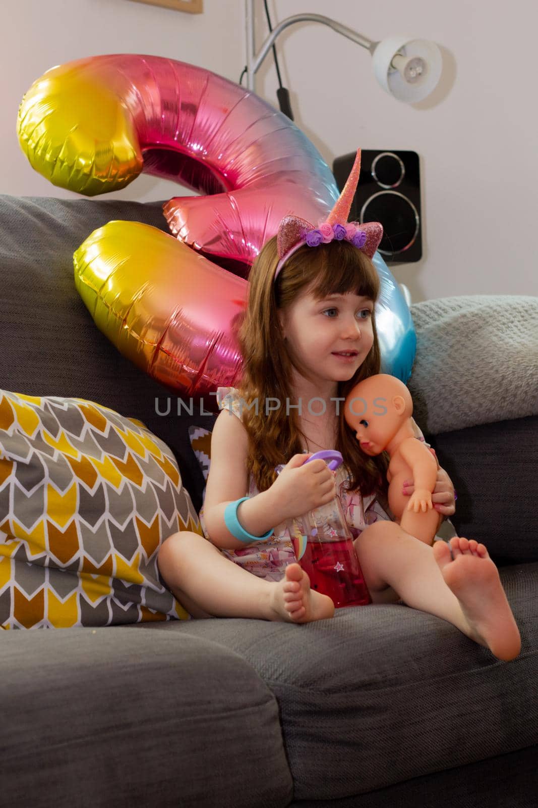 A Cute Baby Girl Sitting On A Sofa With A Colourful Balloon And A Doll by AlbertoPascual