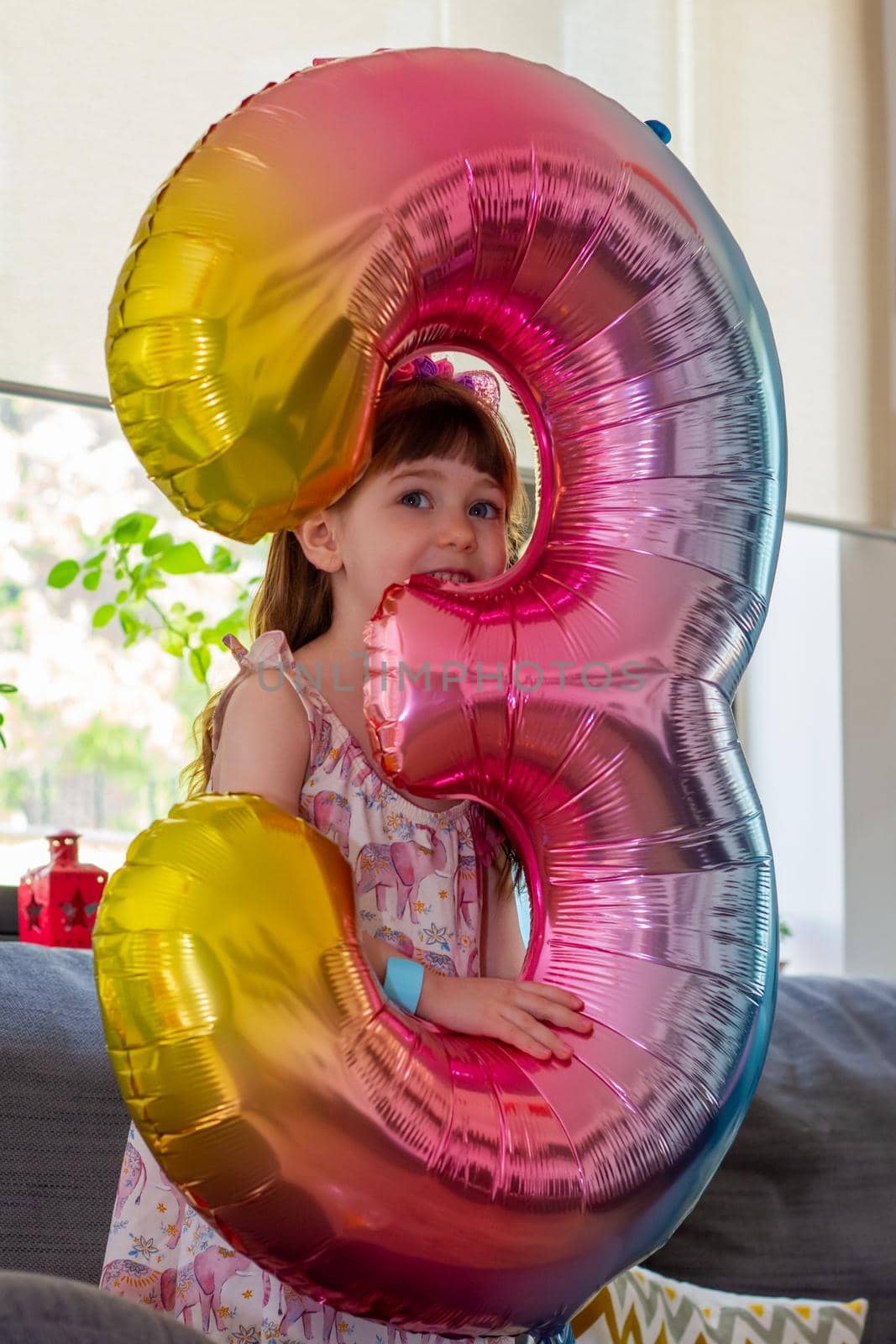 Portrait of a cute, brown-haired girl holding a colourful number three balloon and smiling at camera