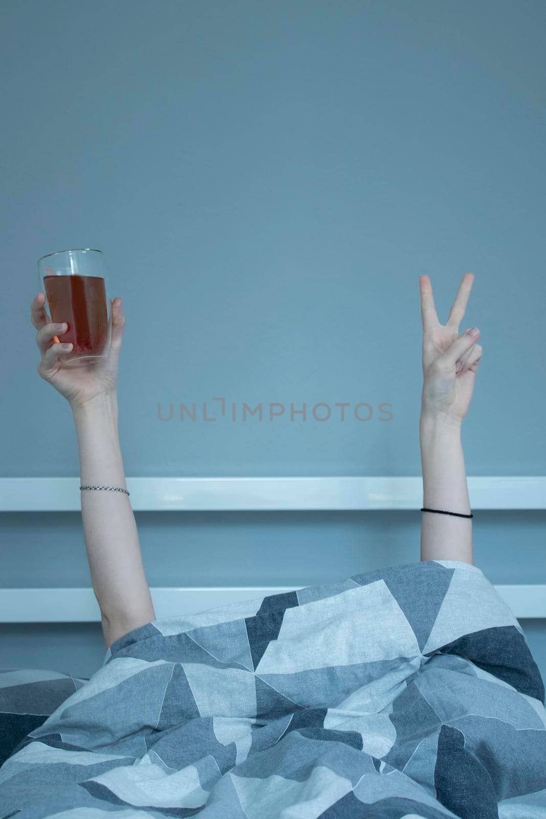 Hands of a girl from under a blanket holding a cup of tea with a double bottom, showing a pease sign with her fingers