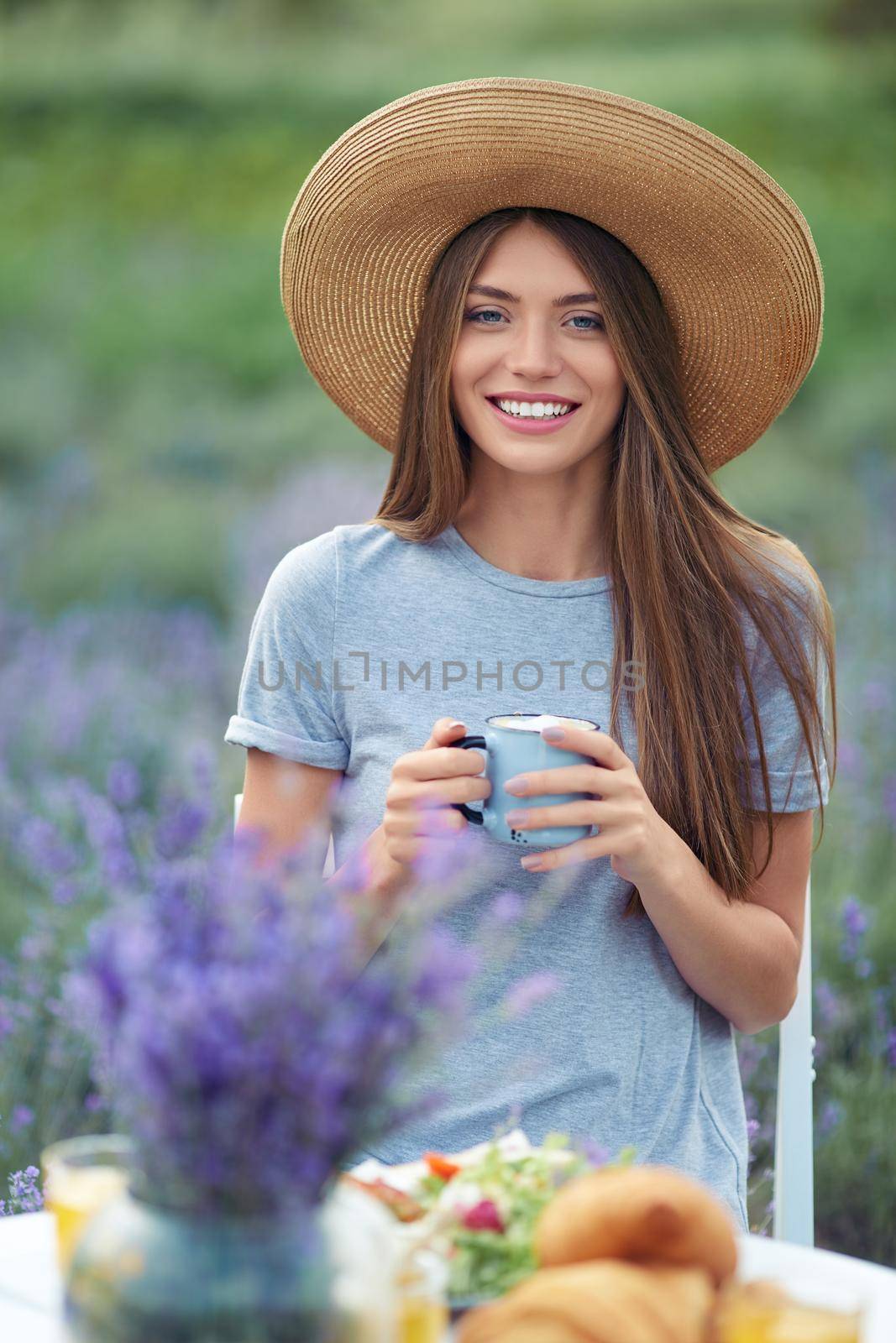Front view of young smiling woman holding cup of coffee, posing at table served with croissants, plate, flowers bouquet and honey jar. Gorgeous girl wearing straw hat sitting in lavender field.