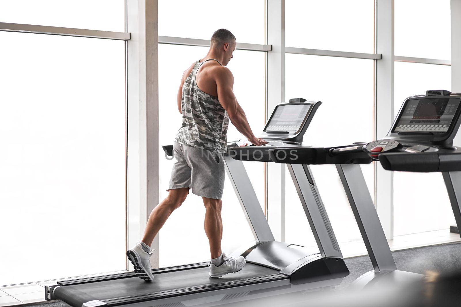 Muscular athletic bodybuilder fitness model running treadmill gym near big window jogging stationary machine do cardio training Weight loss Healthy lifestyle concept. Back view rear full length