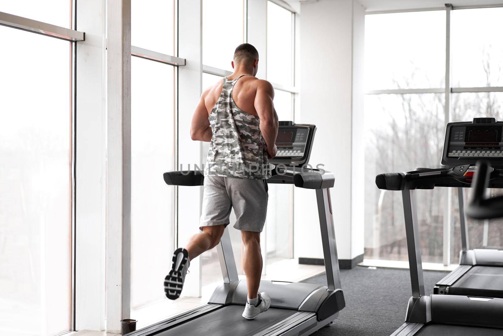 Muscular athletic bodybuilder fitness model running treadmill gym near big window jogging stationary machine do cardio training Weight loss Healthy lifestyle concept. Back view rear full length
