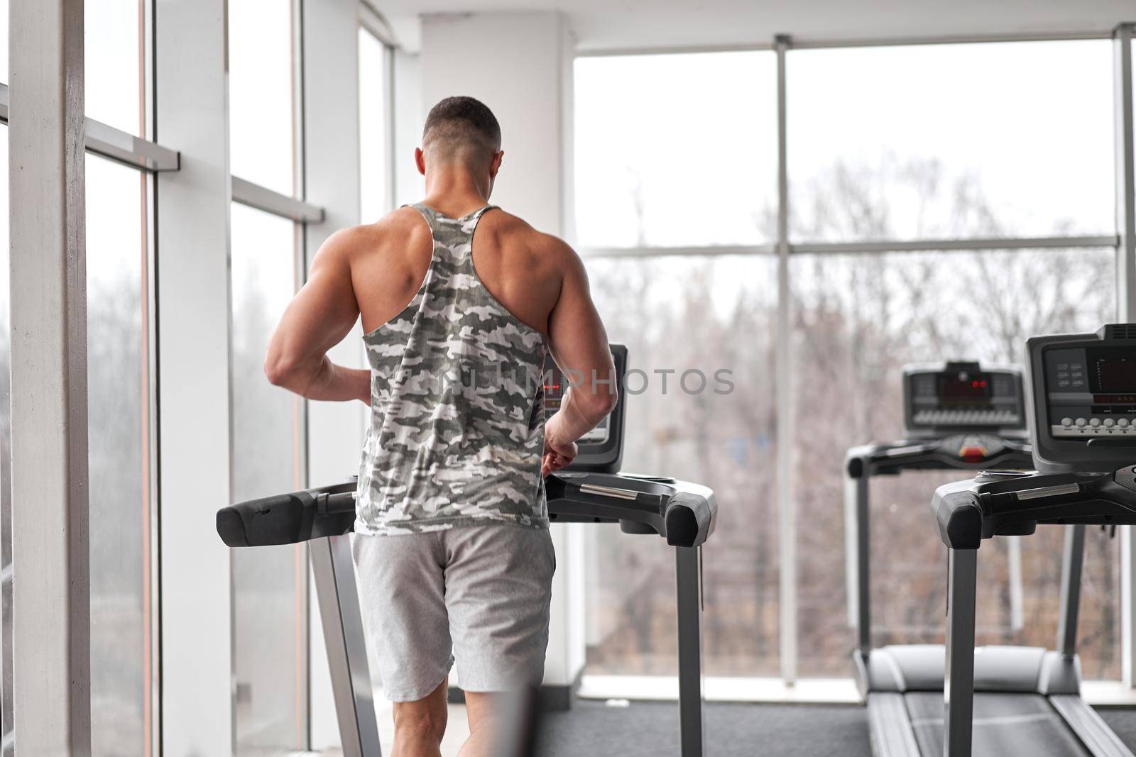 Muscular athletic bodybuilder fitness model running treadmill gym near big window jogging stationary machine do cardio training Weight loss Healthy lifestyle concept. Back view rear