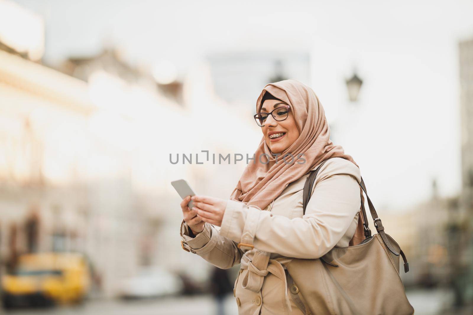 Smiling Muslim woman wearing a hijab and using her smartphone while standing in urban environment.