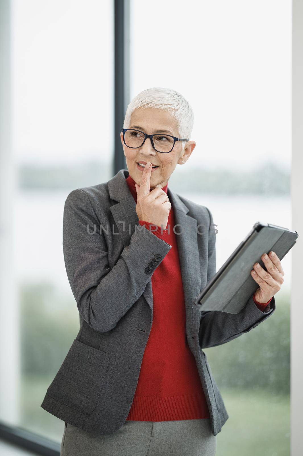 Portrait of a pensive senior businesswoman working on a digital tablet while standing in a modern office near the window.