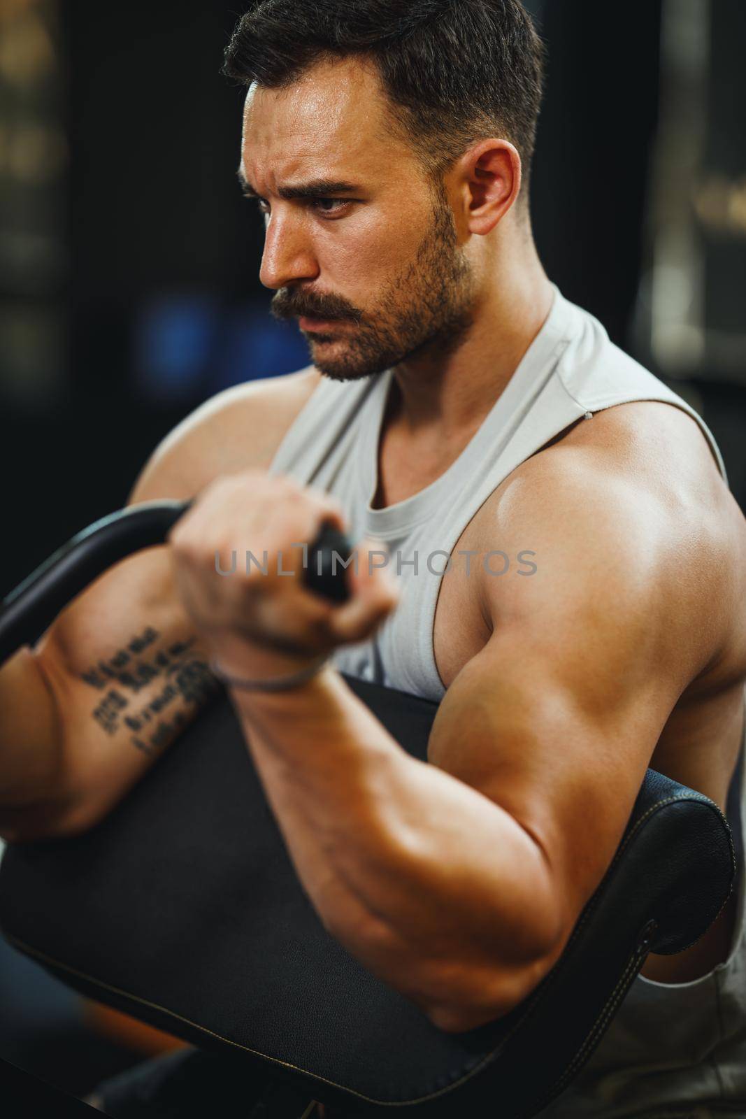 A muscular guy in sportswear working out at the cross training gym. He is pumping up biceps muscule with heavy weight.