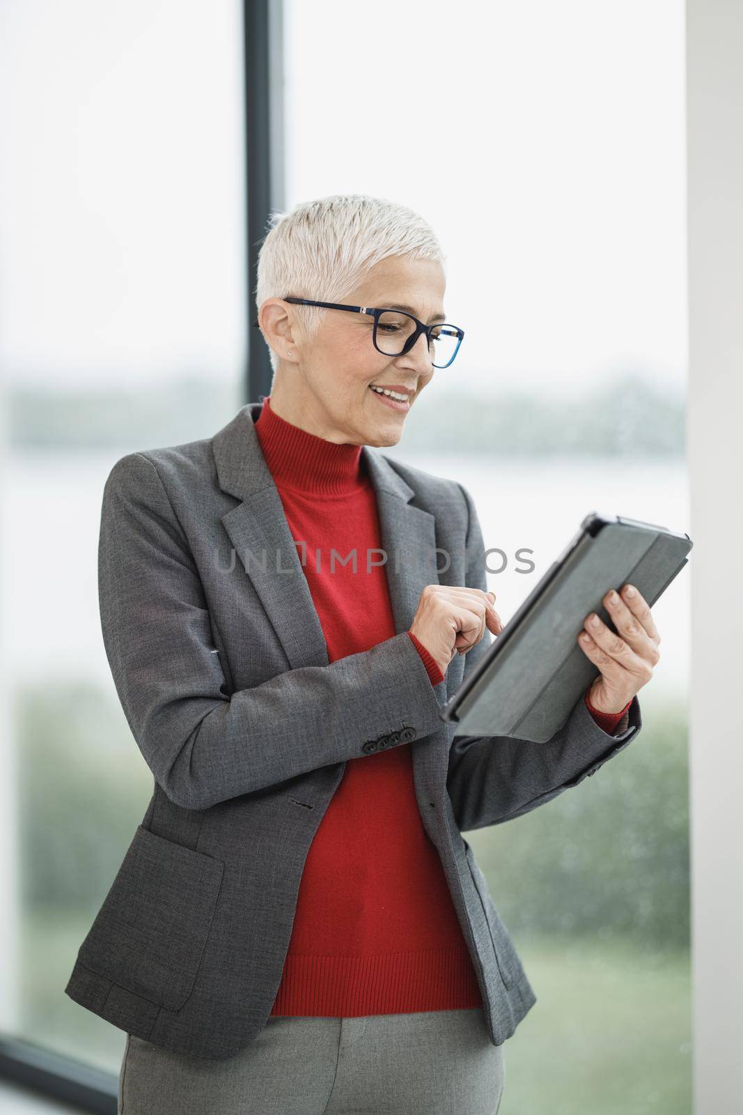 Portrait of a smiling mature businesswoman using app on a digital tablet while standing in a modern office near the window.