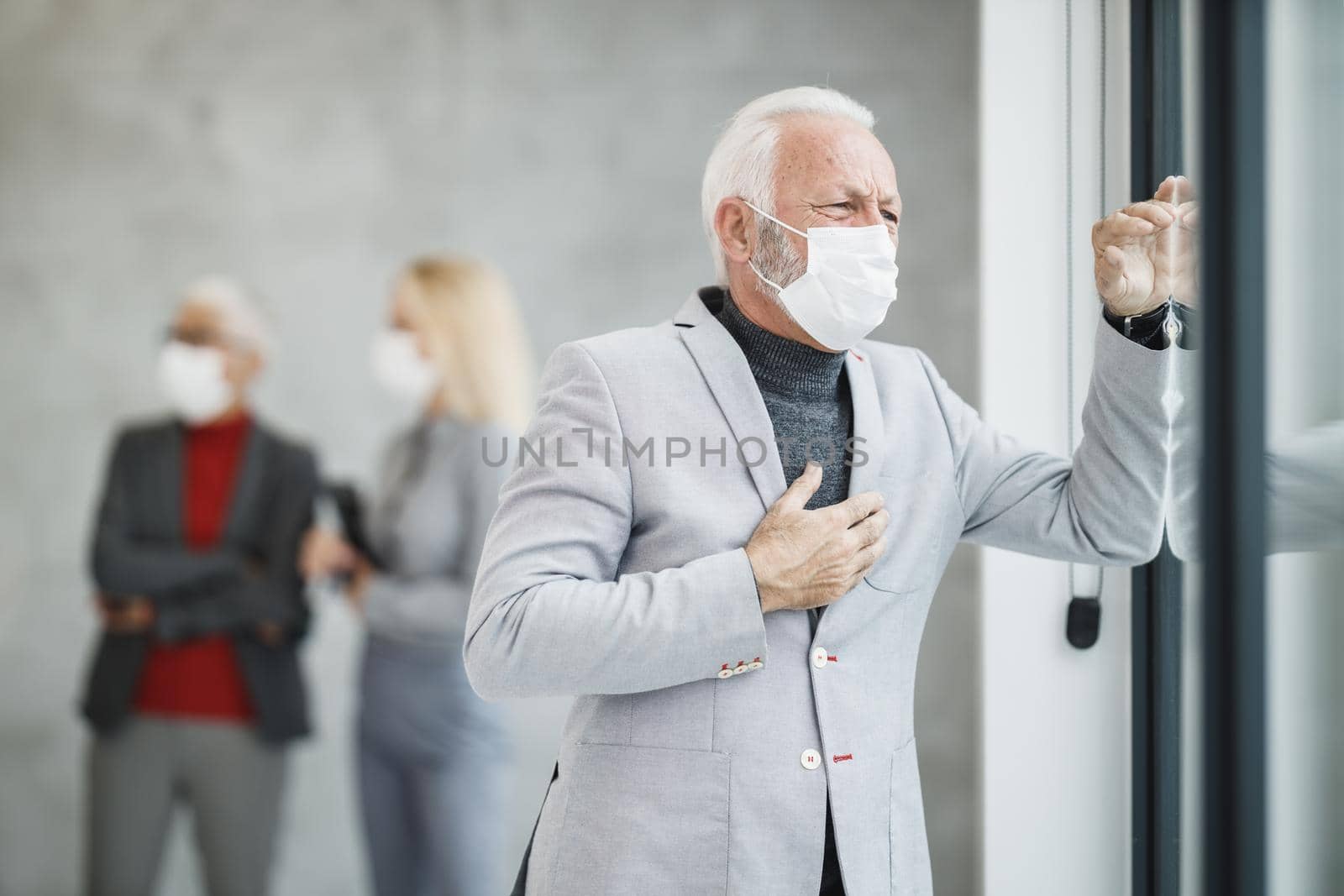 A worried senior businessman with protective mask standing in a modern office and looking out the window during corona virus or COVID-19 pandemic.