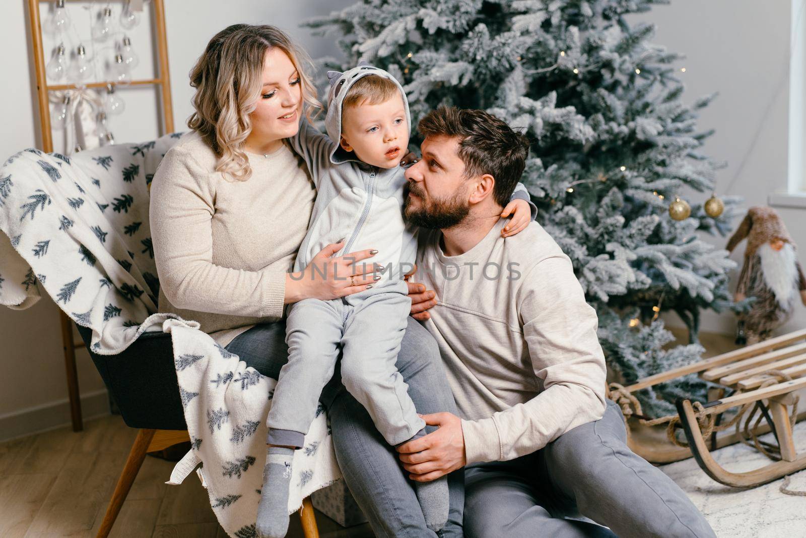 Christmas Family Happiness Portrait of dad, pregnant mom and little son sitting armchair at home near Christmas tree hug smile European young adult family holiday morning