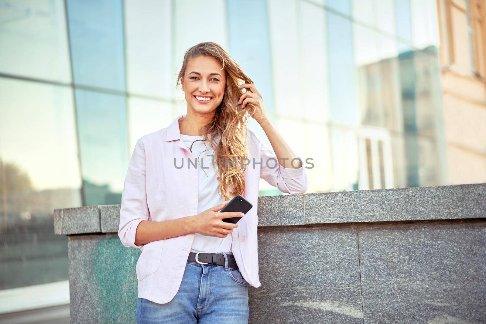 Businesswoman standing summer day near corporate building using smartphone teeth smiling Business person Outdoors Successful european caucasian woman freelancer dressed white shirt pink jacket