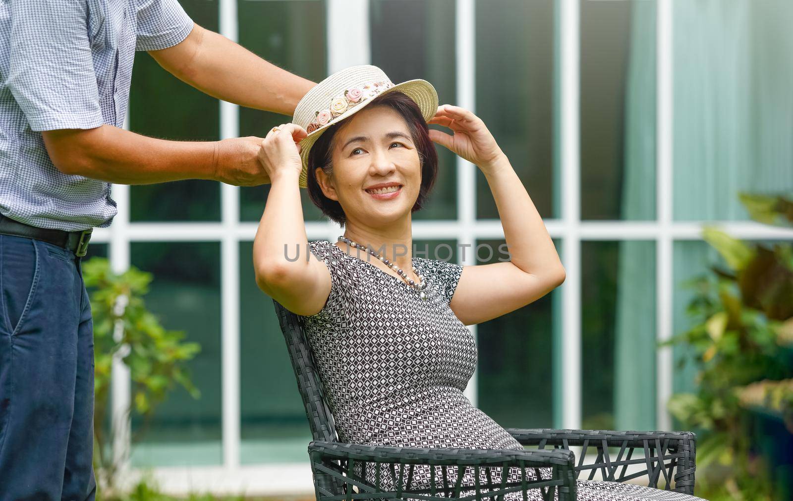 Asian middle aged couple sitting relax in backyard and putting on a hat to his wife