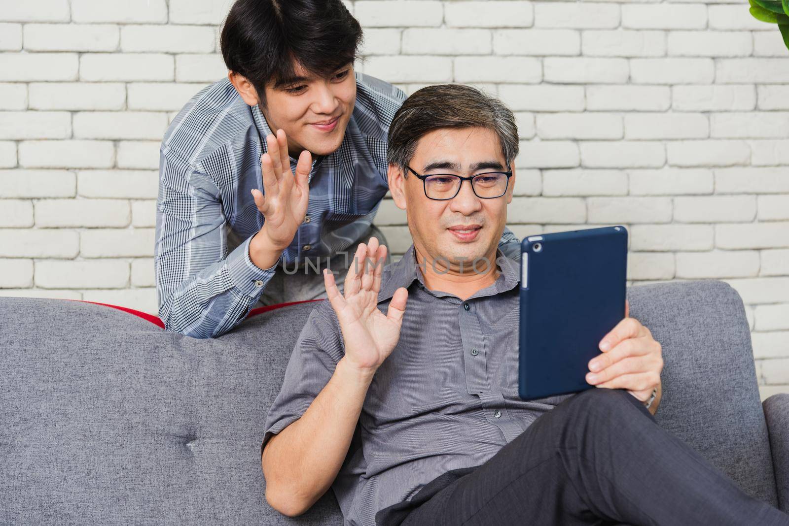 Asian senior businessman with digital tablet discuss together with young team in office. Father man and his son sit on sofa talking chatting on video call conference on tablet in living room at home