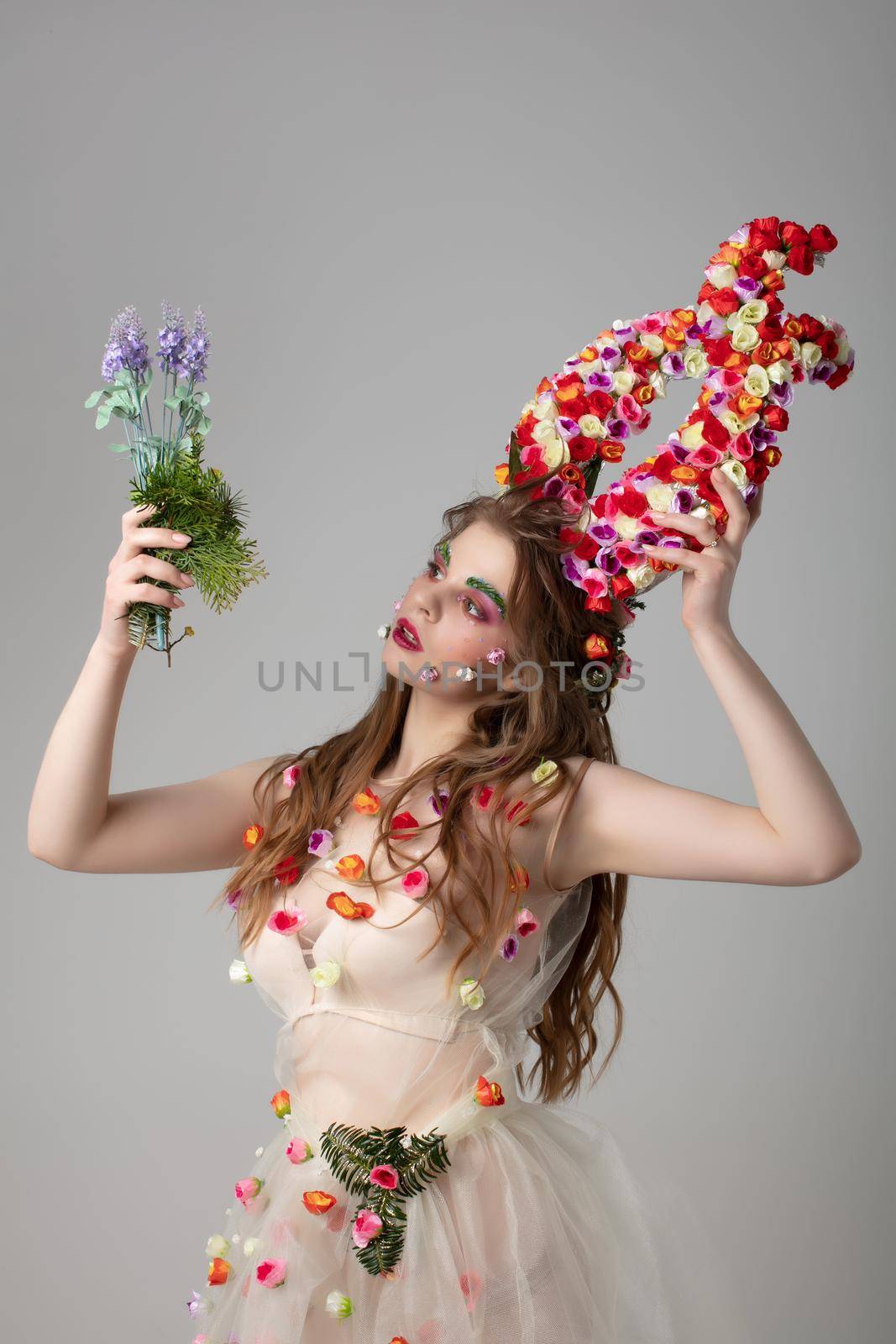 Fairy-tale girl model with floral horns and a forest bouquet on a studio background. Maleficent. Spring or summer beauty. Woman in the image of a flower. Goddess Flora