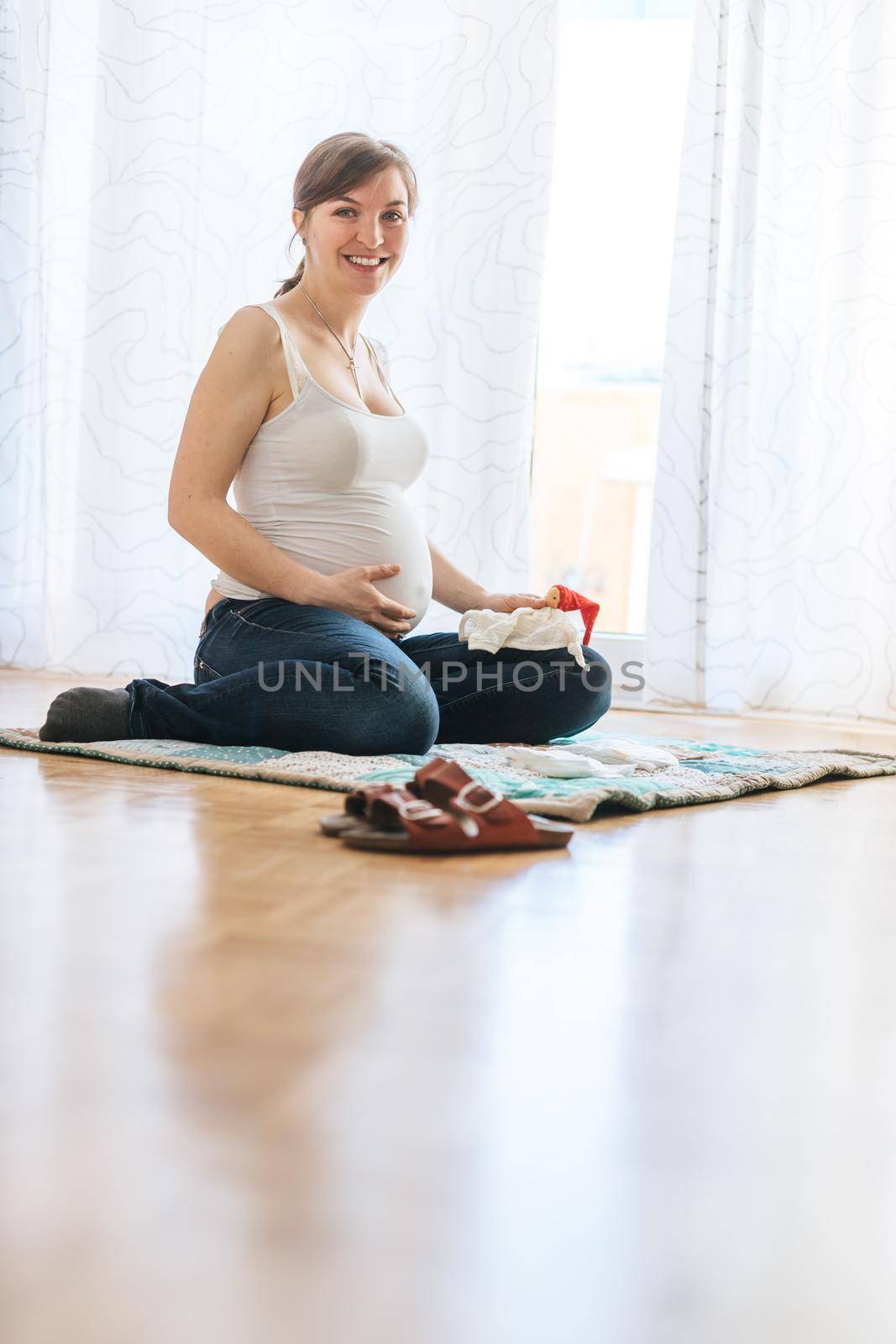 Happy Caucasian pregnant mother is sitting on the floor, touching her tummy, blue jeans